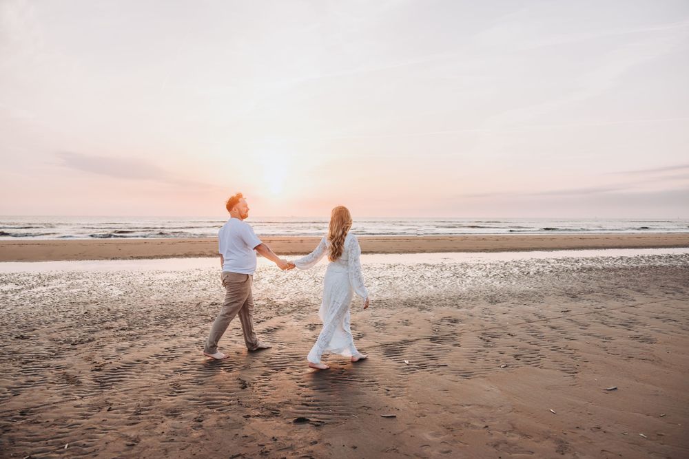 Warm and natural edit - preregnant couple holding hands walking on a sandy beach at sunset, woman in white dress, romantic seaside stroll.