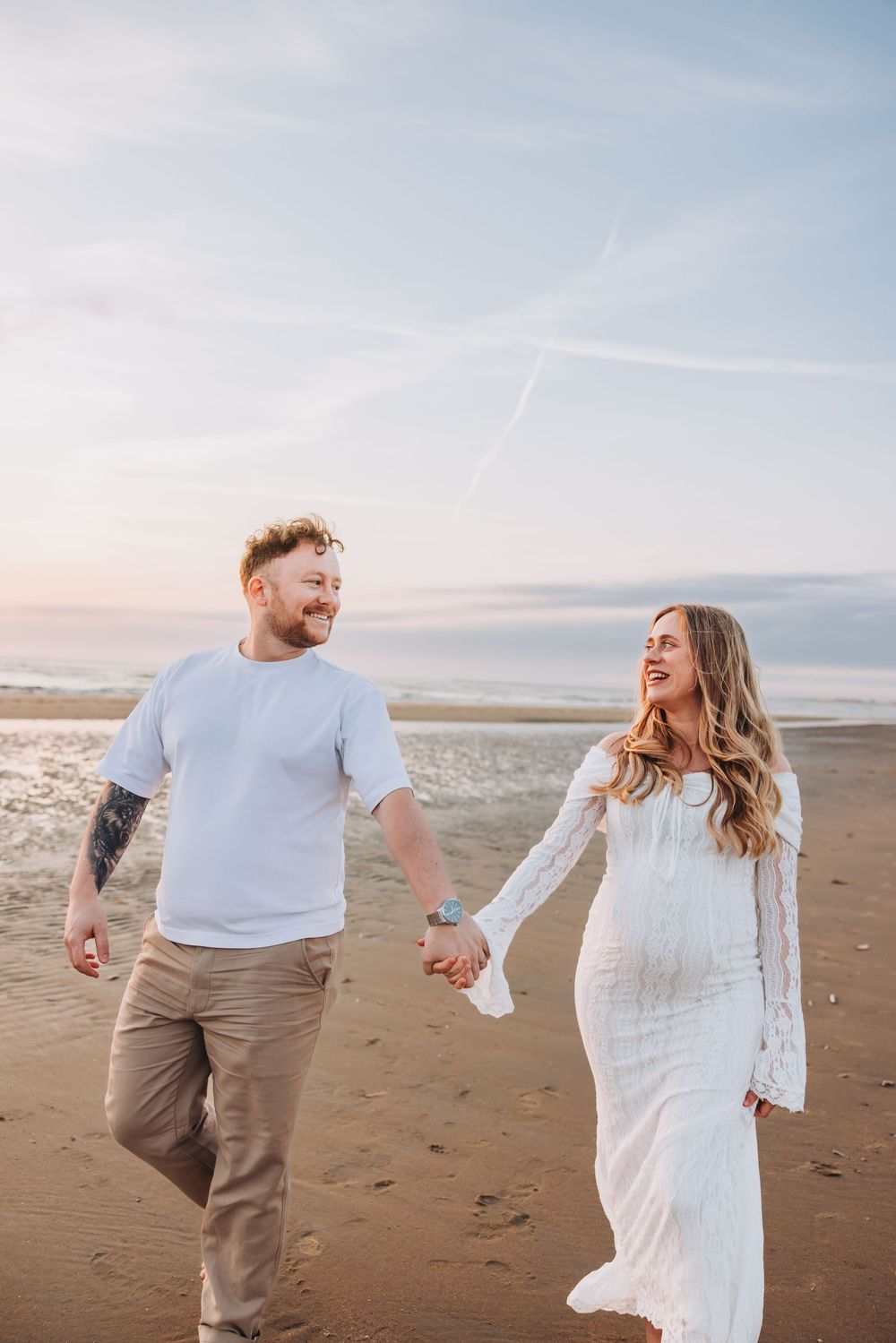 A happy pregnant couple holds hands and walks along a sandy beach at sunset, the woman wearing a white lace maternity dress.