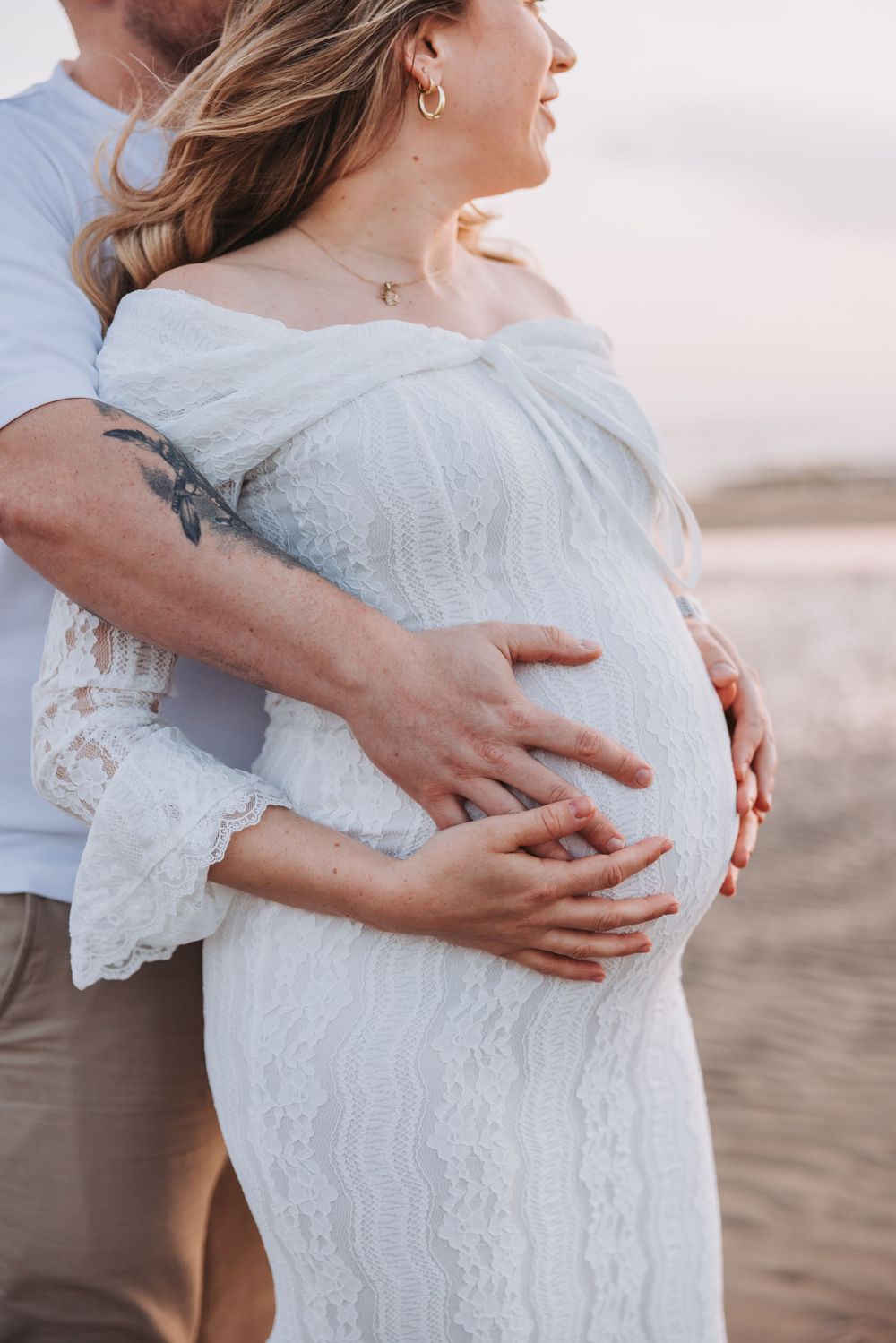 Pregnant woman in white lace dress with partner embracing her baby bump on a beach at sunset.