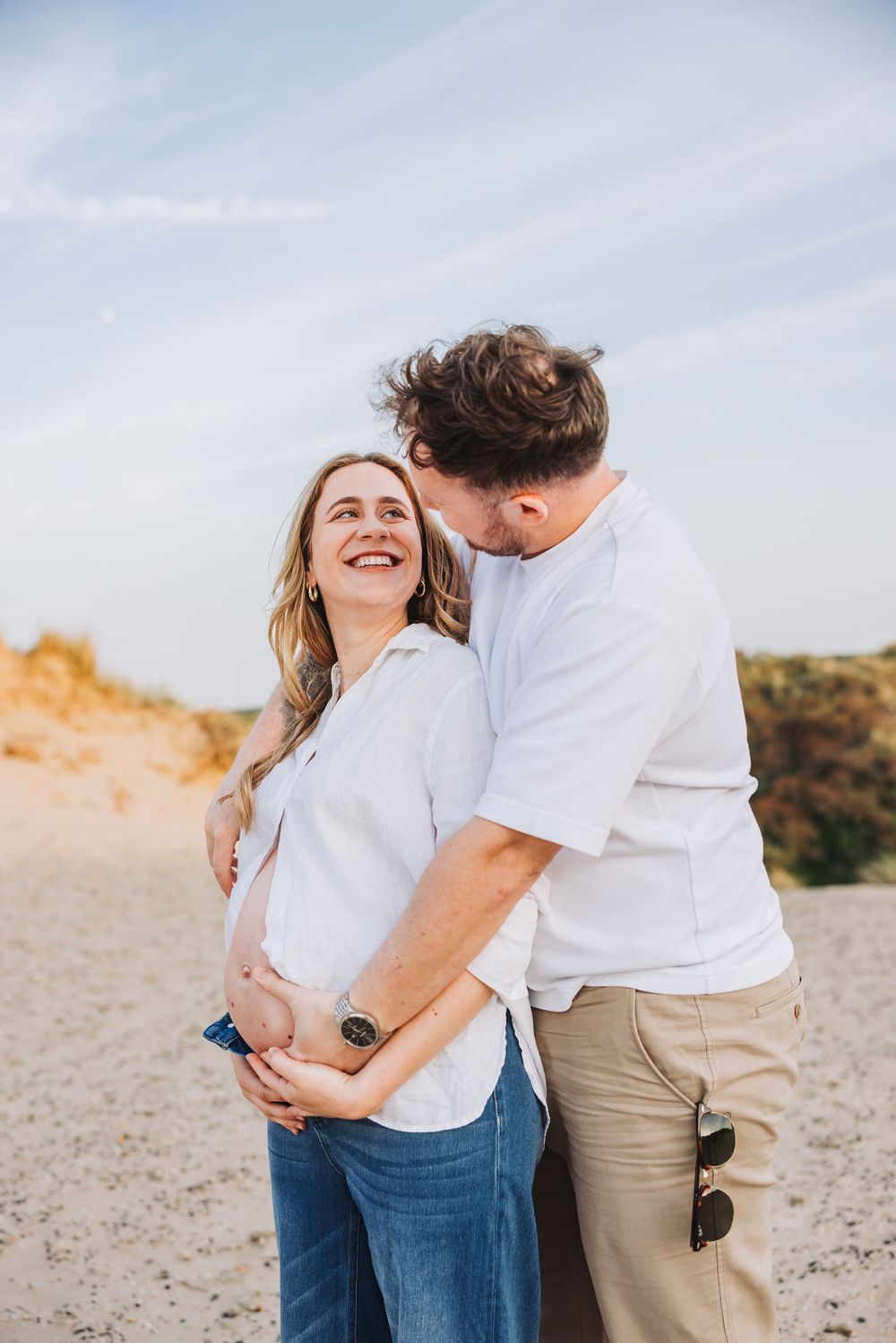 Pregnant woman laughing joyfully as man kisses her cheek amidst sunny dunes during summer beach photoshoot at Bloemendaal.