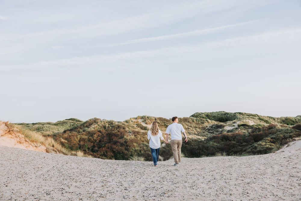 Couple holding hands walking on a sandy beach with green dunes on the Dutch coast under a clear sky.