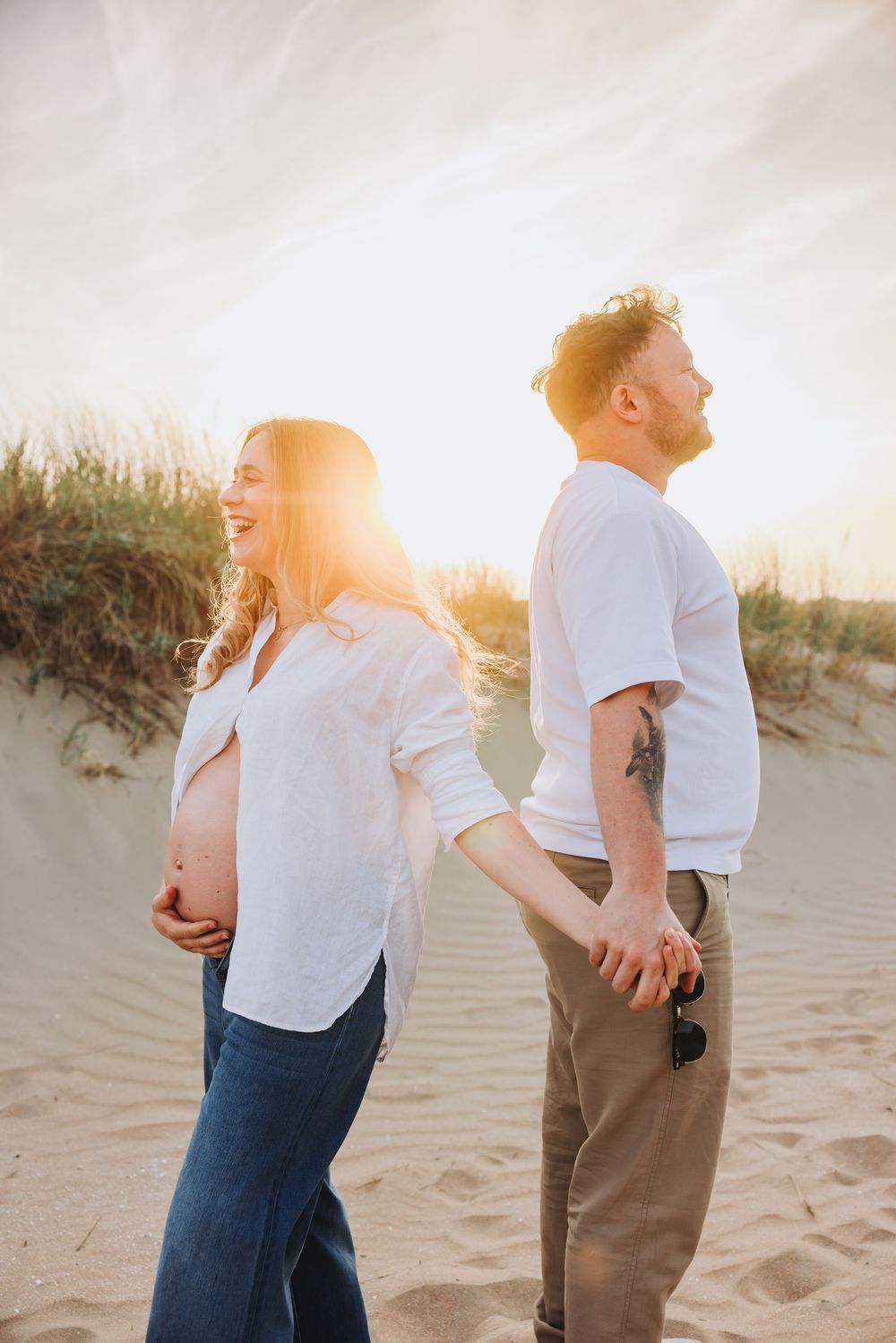 Pregnant couple holding hands back to back on sandy dunes at golden sunset, woman in white shirt laughing joyfully. Warm golden editing