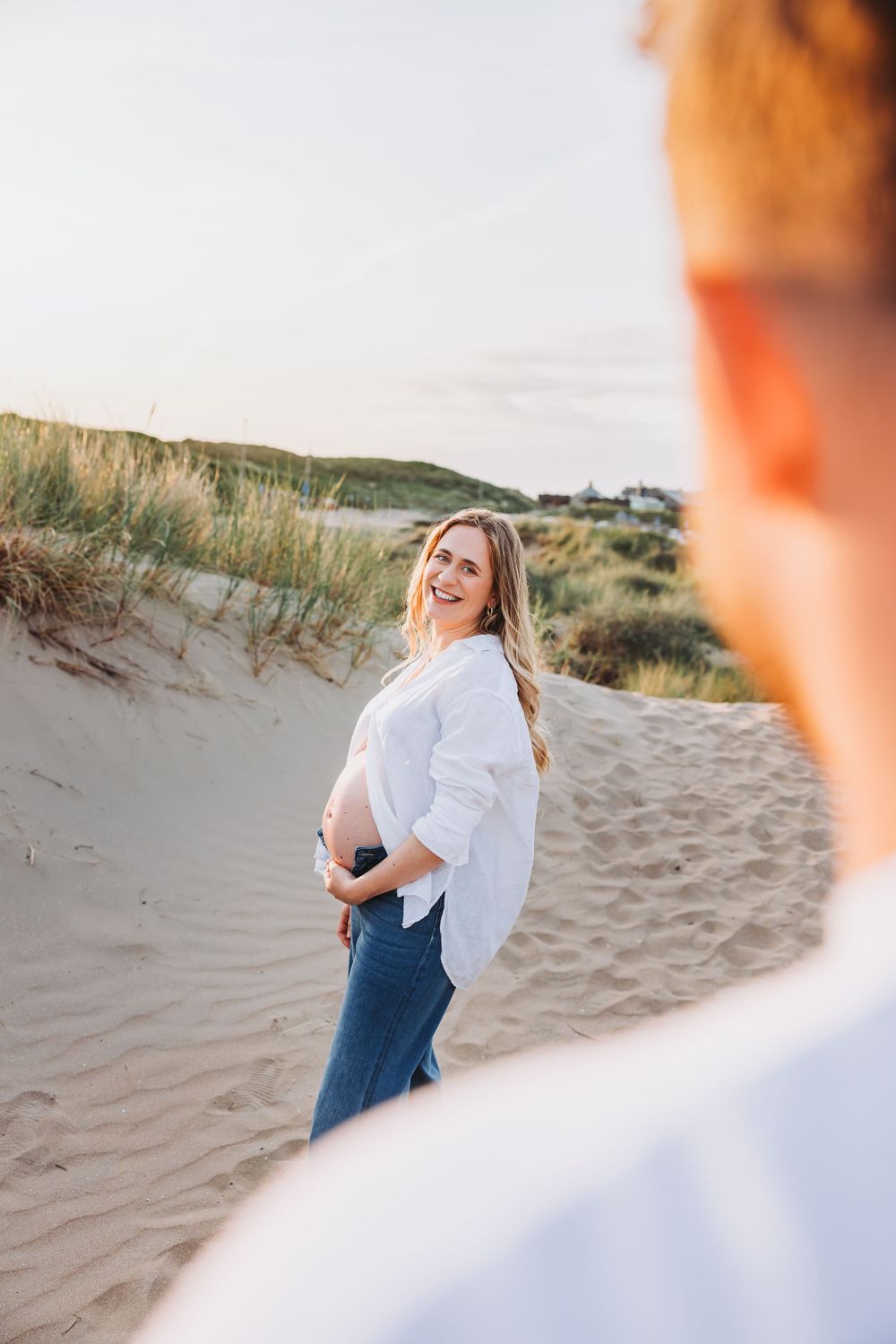 Over the shoulder shot of smiling pregnant woman on beach dunes, warm golden hour edit.