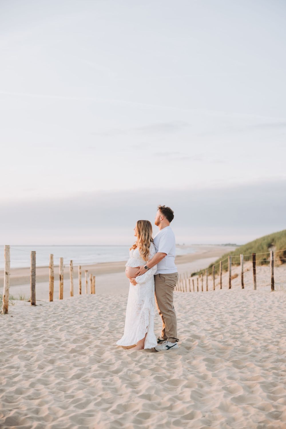 Pregnant couple embracing on sandy path to Bloemendaal beach at sunset, woman in white dress, man in white shirt, khaki pants, natural edit.