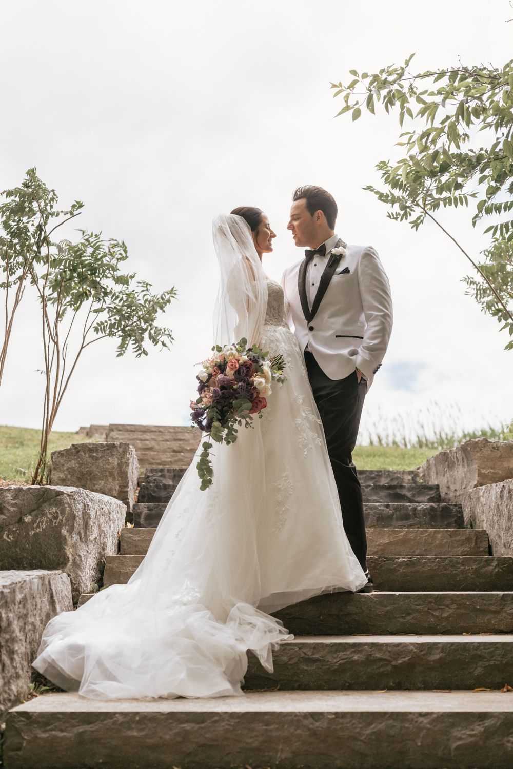 Bride and groom share a romantic moment on stone steps, bride holding a bouquet in a white gown with veil.