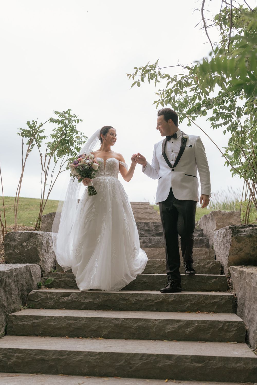 Bride and groom hold hands on stone steps, she in a lace gown with veil and bouquet, he in a white tuxedo jacket.