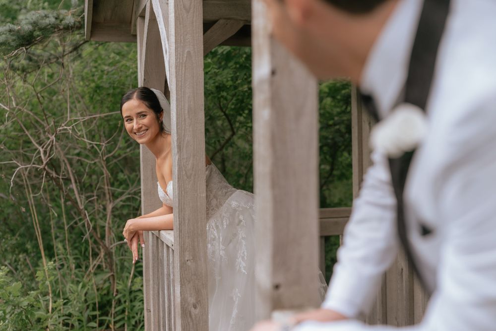 Bride in white gown leans on wooden balcony smiling at groom in white suit outdoors.