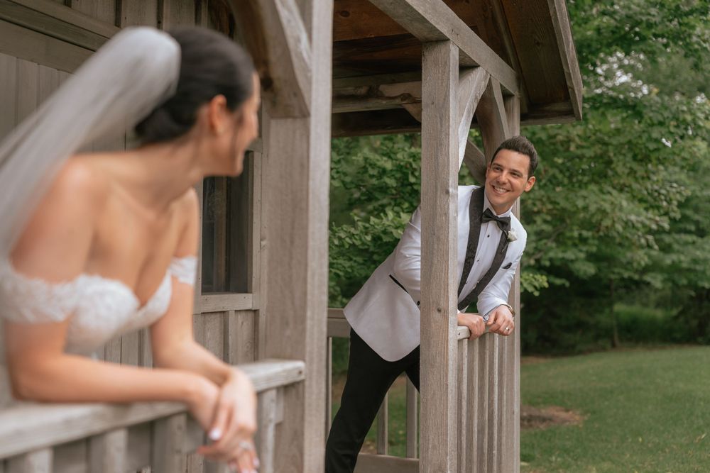 Groom in white tuxedo jacket smiles at bride in strapless wedding gown on rustic wooden porch during first look.