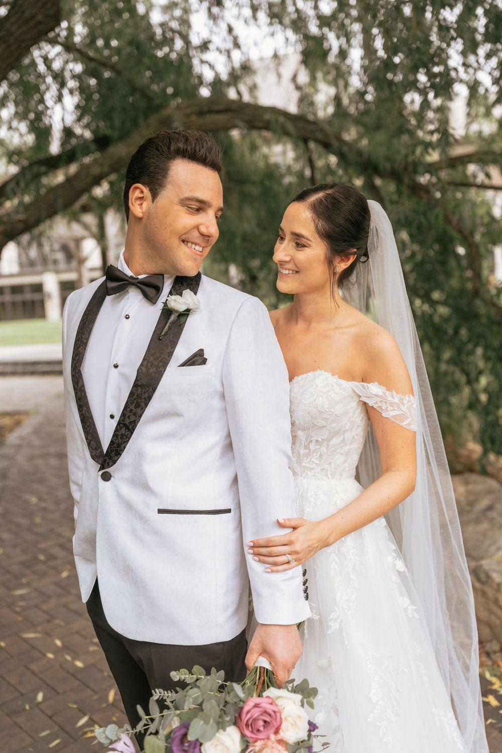 Bride and groom smile at each other outdoors, she holds a floral bouquet, wearing a lace gown and veil, he wears a white tuxedo.