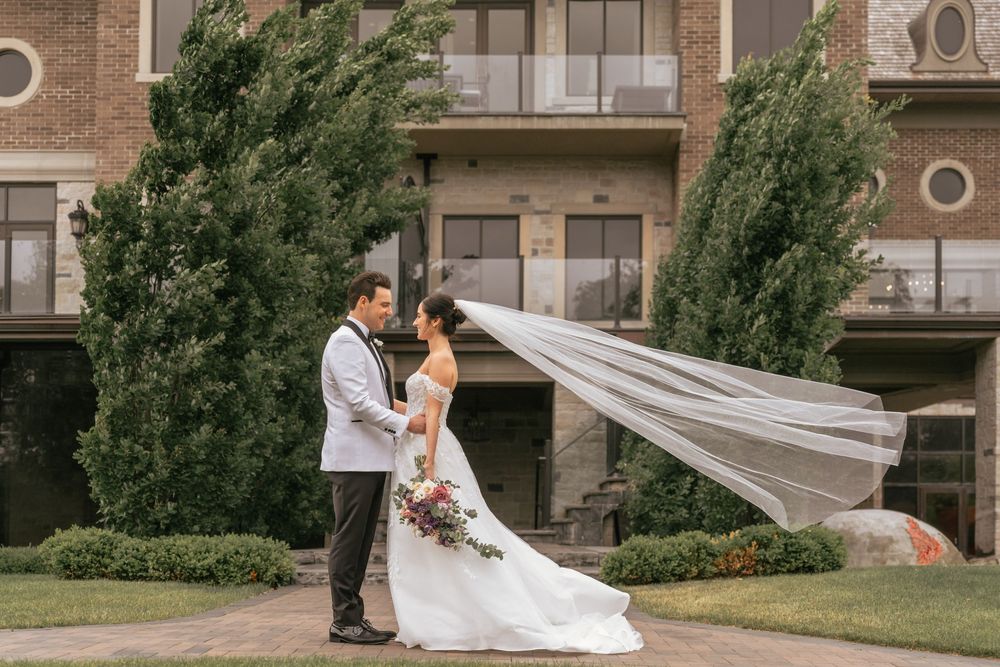 Bride and groom share a romantic moment outdoors, her veil flowing in the wind, with a brick building and lush greenery behind.