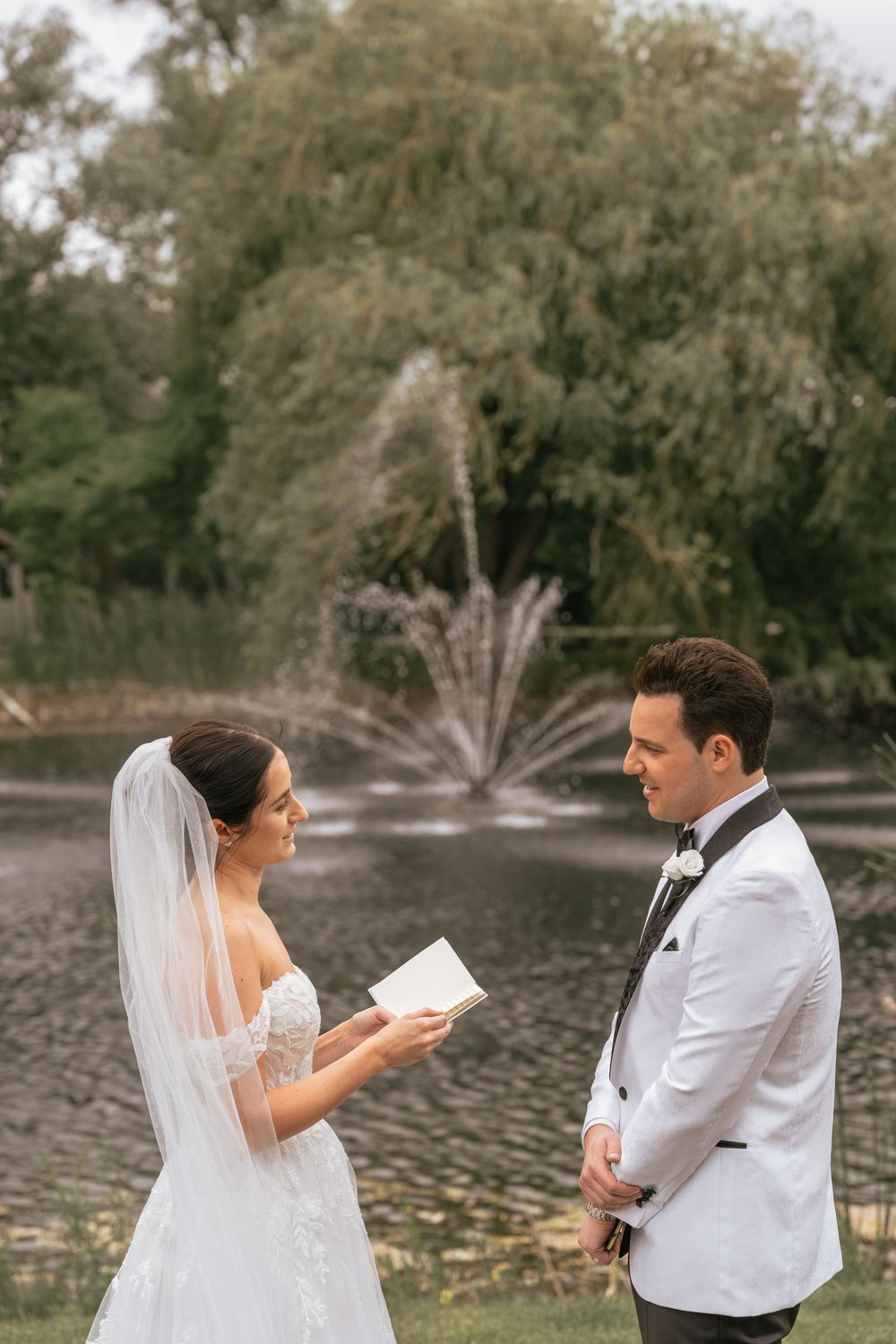 Bride reads wedding vows to groom in white tuxedo by a fountain and pond surrounded by lush greenery.