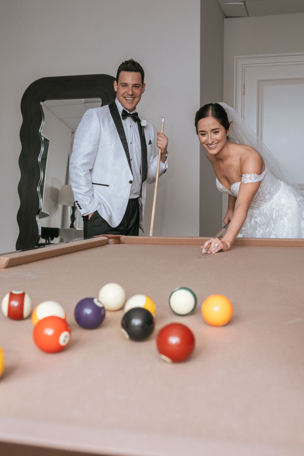 Bride and groom playing pool on their wedding day, smiling at the billiard table in wedding attire.