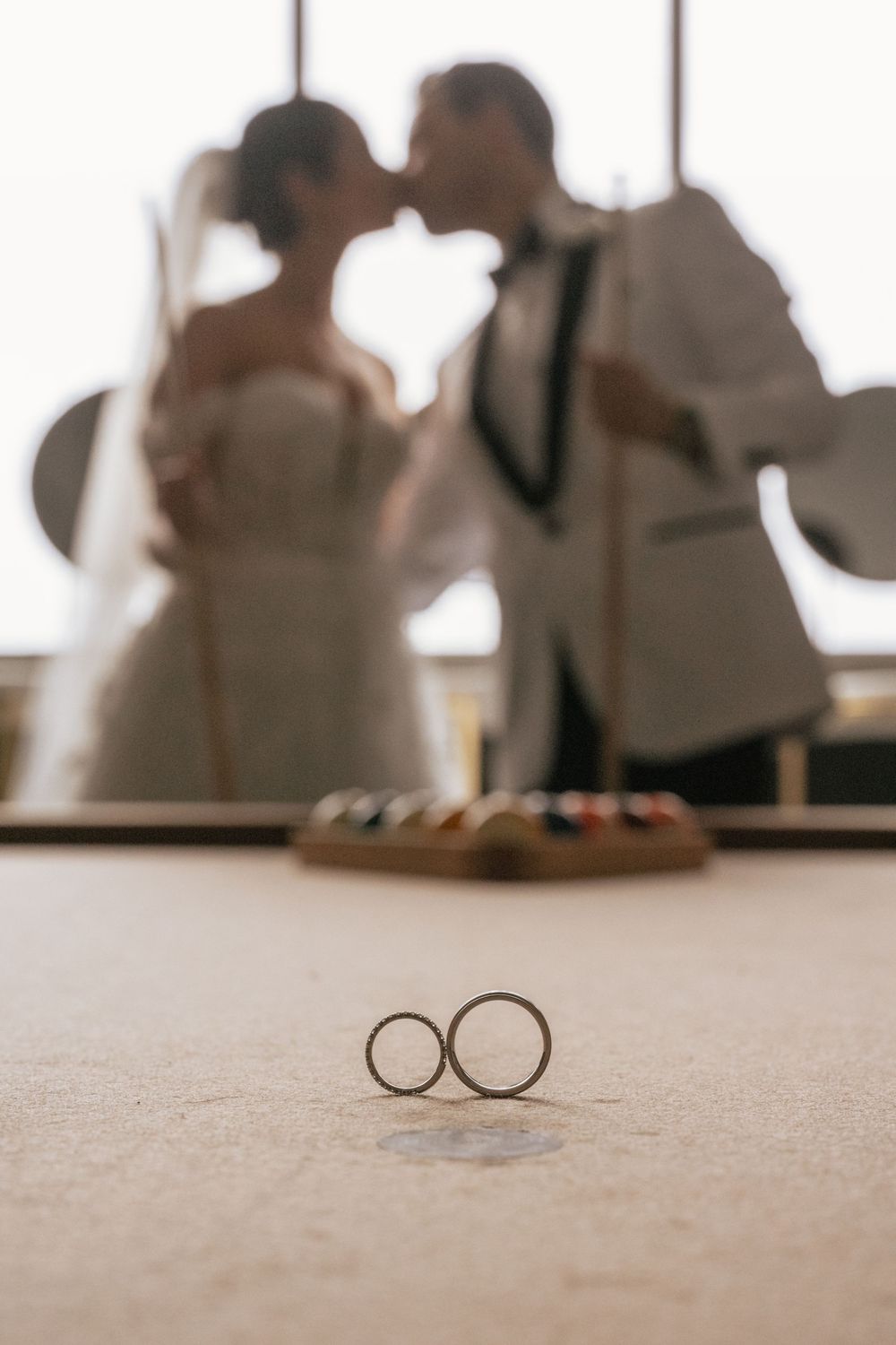 Two wedding rings in focus on a table with a blurred bride and groom kissing in the background.