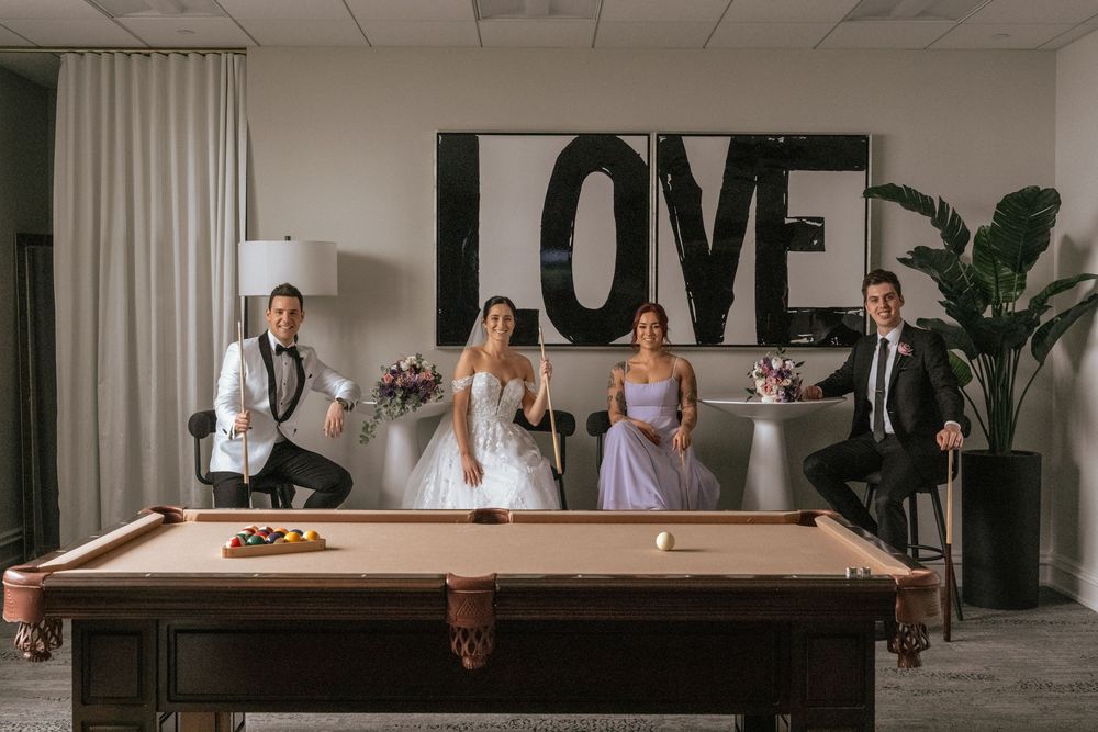 Wedding party poses around a pool table with a large LOVE sign backdrop in a modern venue.