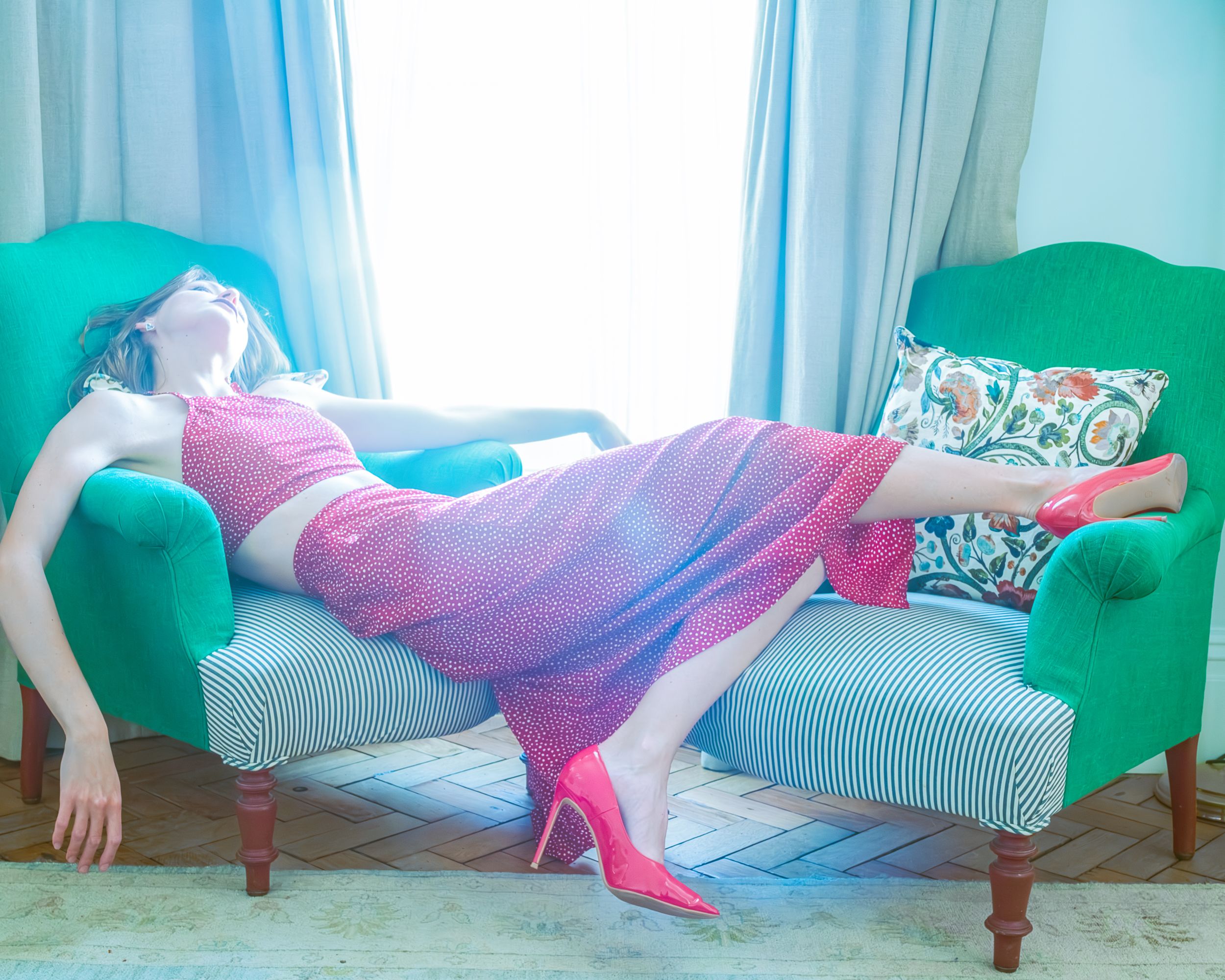 Model reclining on a teal vintage sofa wearing a flowing pink gown, colorful editorial fashion photo