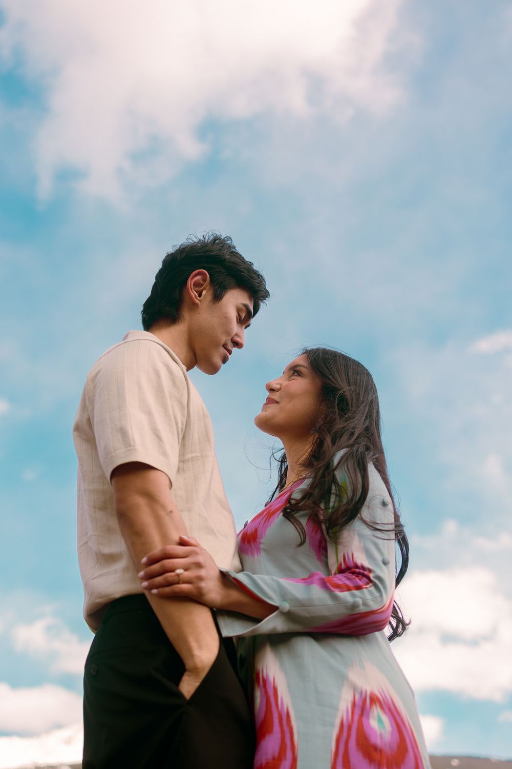 A young couple embraces outdoors, gazing into each other's eyes against a bright blue cloudy sky.