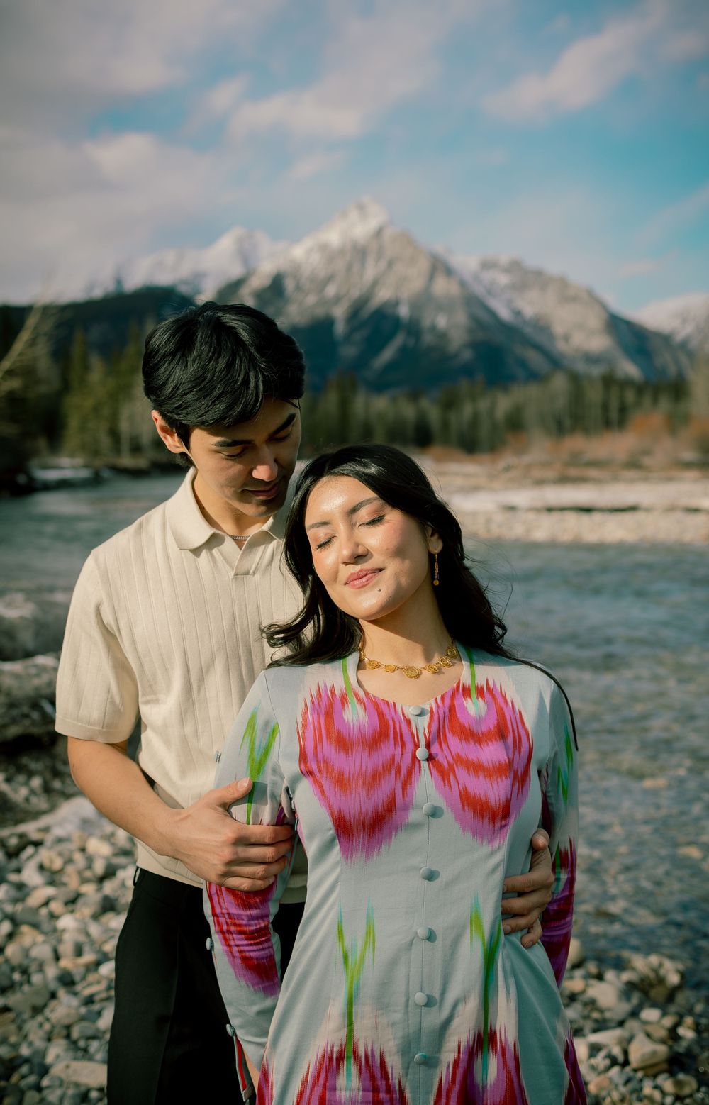Couple embracing near a rocky river with snow-capped mountains, woman in colorful ikat dress, man in cream shirt.