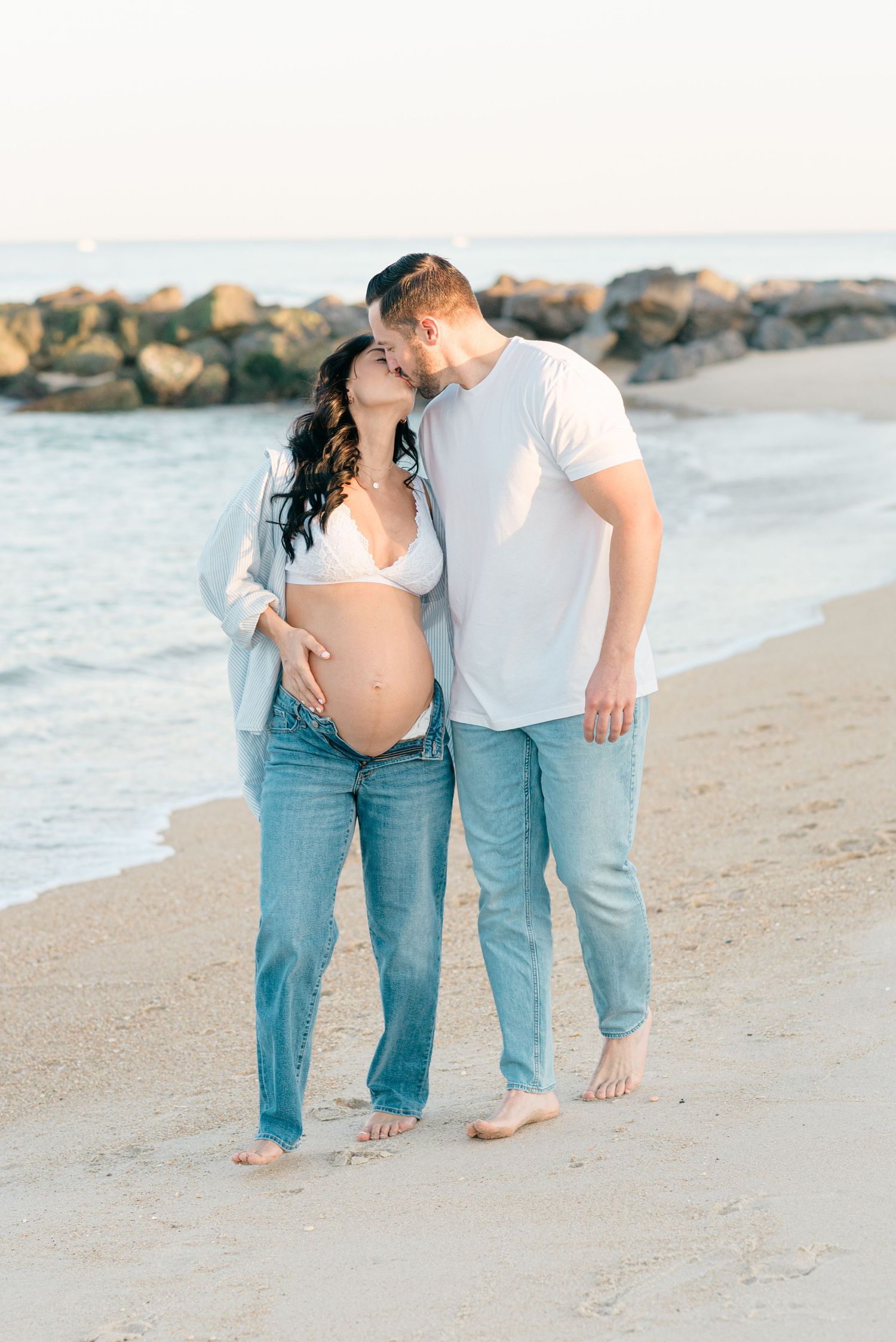 Maternity photography at the Jersey Shore with an expectant mother wearing a white bralette and jeans kisses her husband.