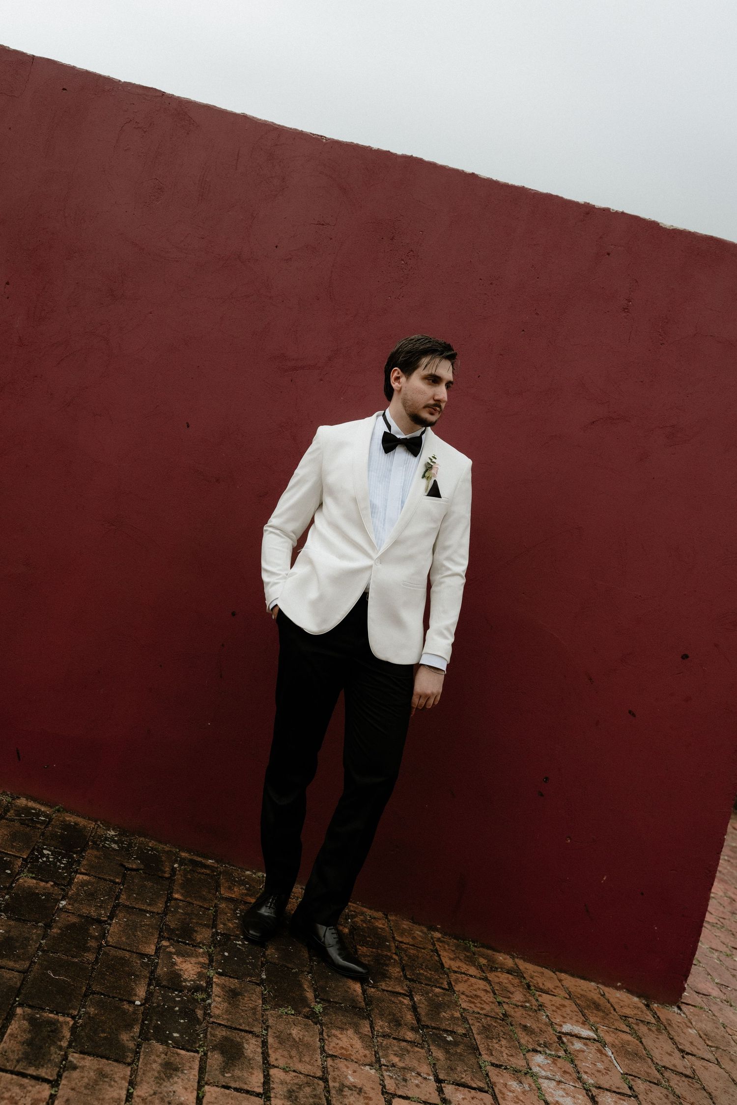 Groom in white tuxedo jacket and black bow tie poses against a deep red wall on brick flooring.