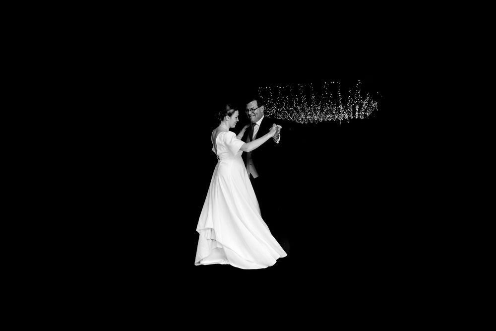 Bride and groom share first dance in elegant black and white wedding photo with chandelier backdrop.