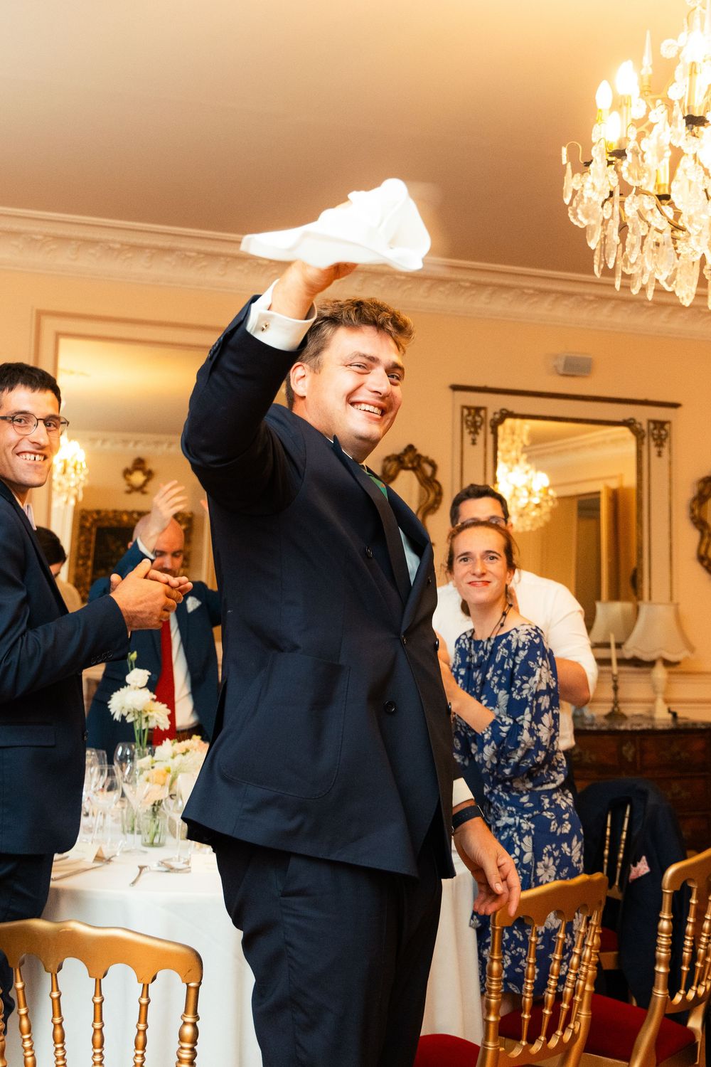 Man in navy suit raises white napkin at elegant wedding reception with chandelier and guests.