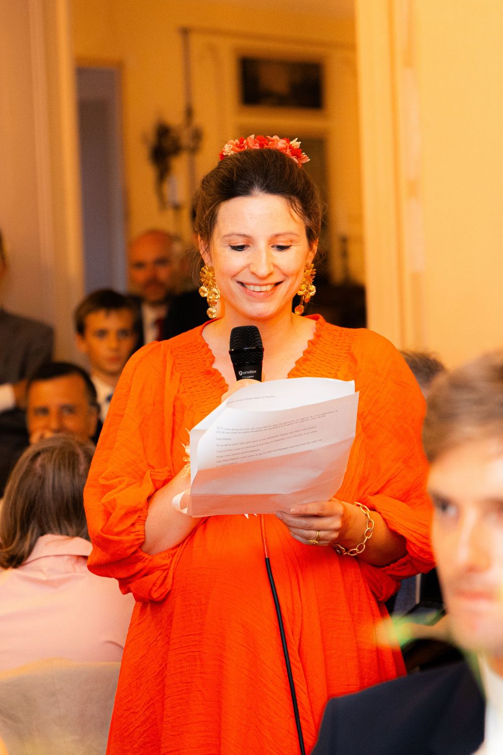 Woman in orange dress smiling while giving a speech at an event, holding papers and a microphone.