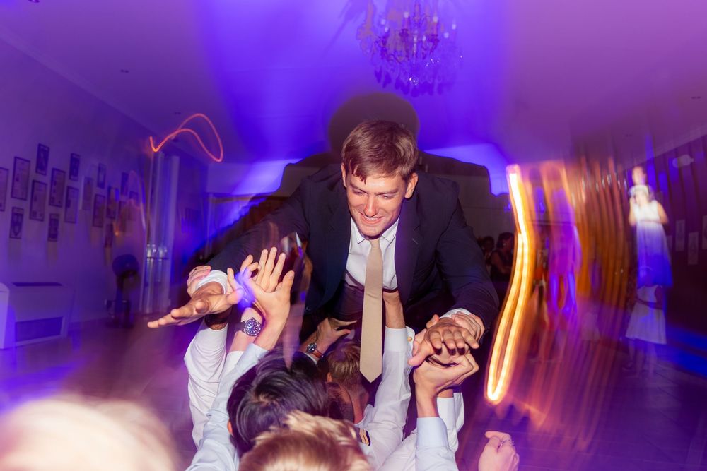 Groom crowd surfing at wedding reception with purple lighting and guests cheering below.