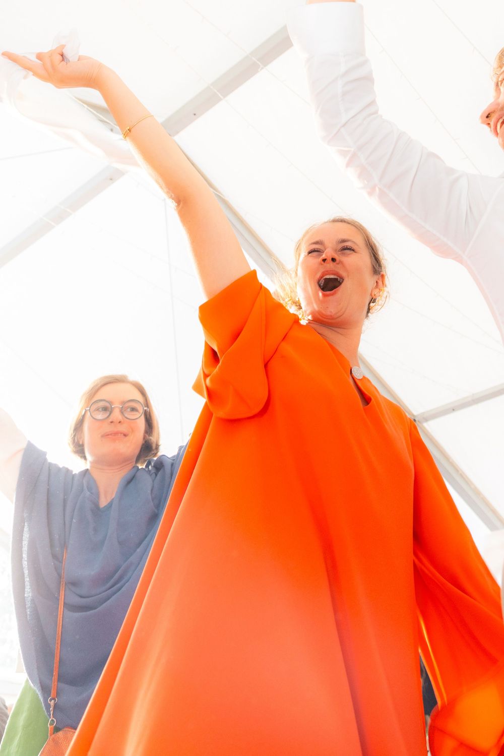 Two women celebrating with raised arms, one in vibrant orange coat, photographed from below at an outdoor event.