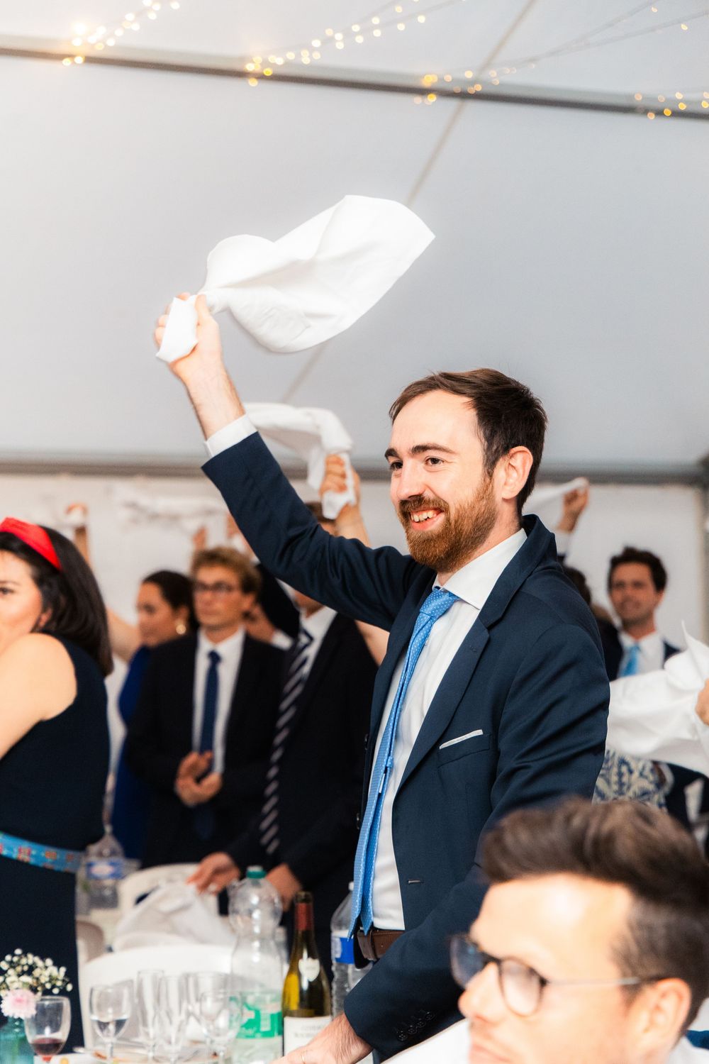 Man in navy suit waving white napkin at wedding reception celebration in marquee tent.
