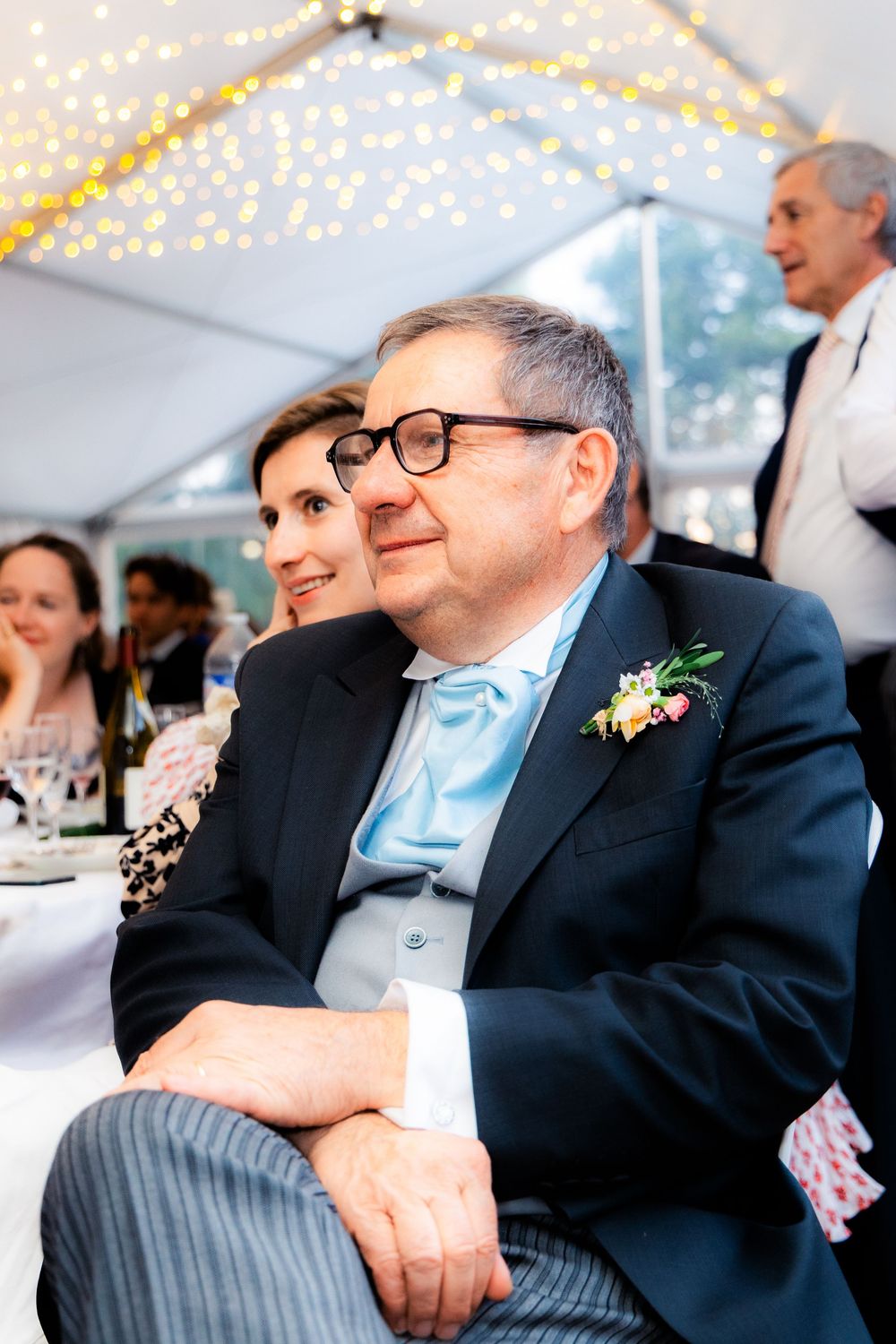 Elderly man in suit with boutonniere and blue tie sits at wedding reception under string lights.