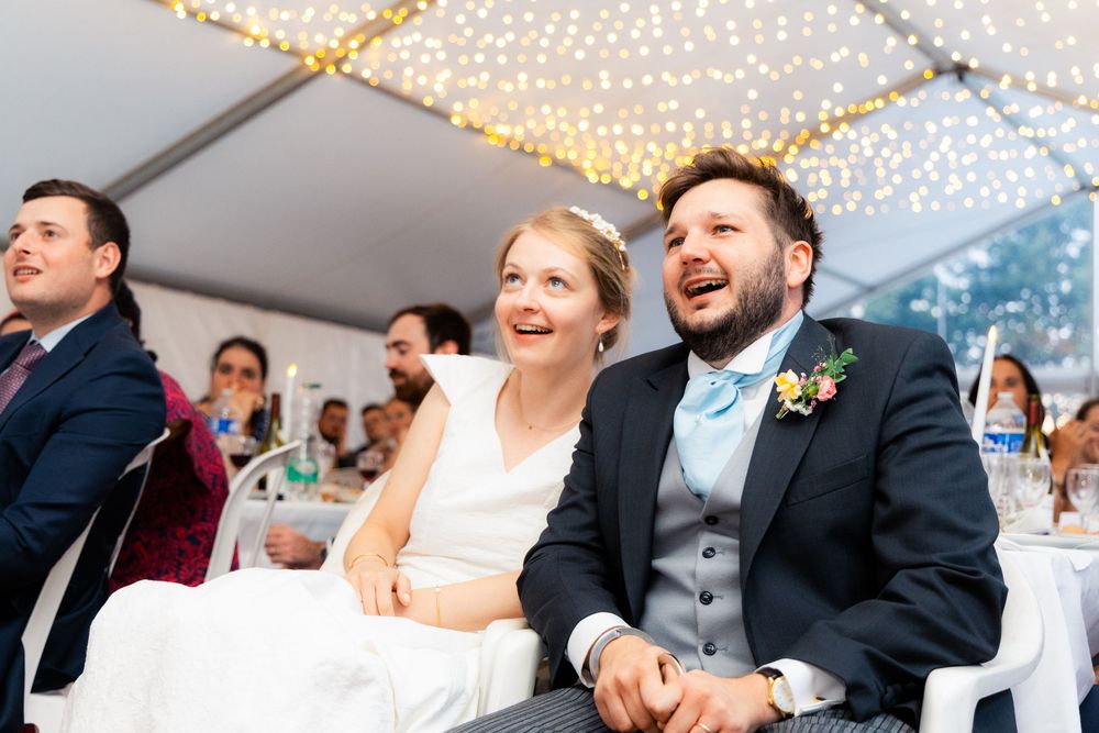 Bride and groom laughing joyfully at wedding reception under fairy lights in elegant tent venue.