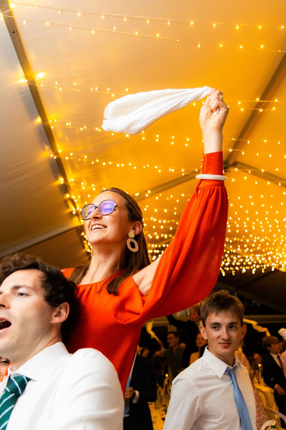 Woman in red dress waving white napkin at a wedding reception under warm string lights.