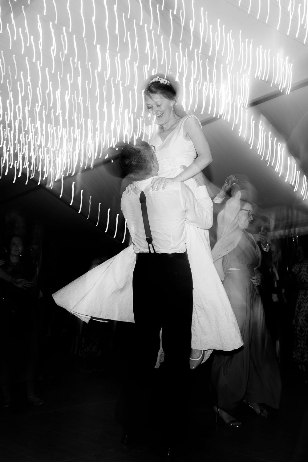 Groom lifting bride in white gown during first dance at wedding reception with sparkling lights backdrop.