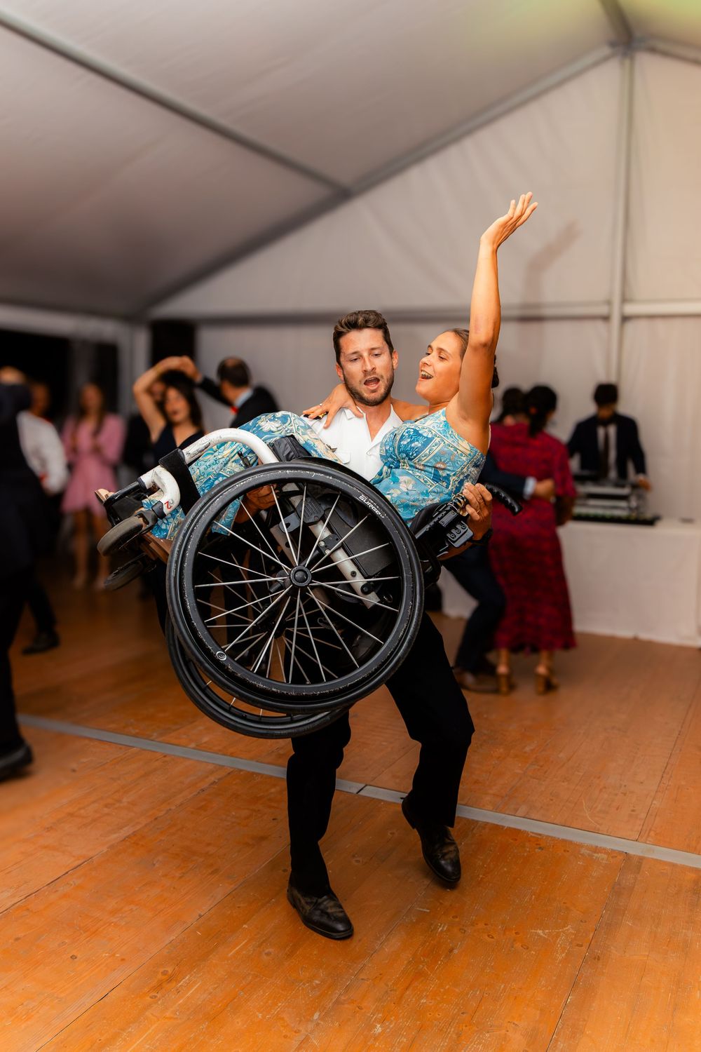 Man carrying woman in wheelchair on dance floor at wedding reception celebration.