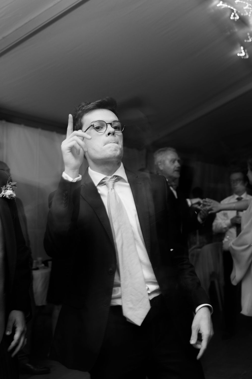 Man in suit and glasses points upward while dancing at a formal wedding reception event.
