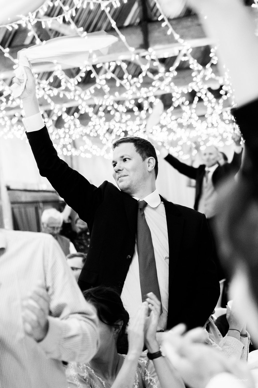 Groom raises hand joyfully at wedding reception with twinkling fairy lights in black and white photo.