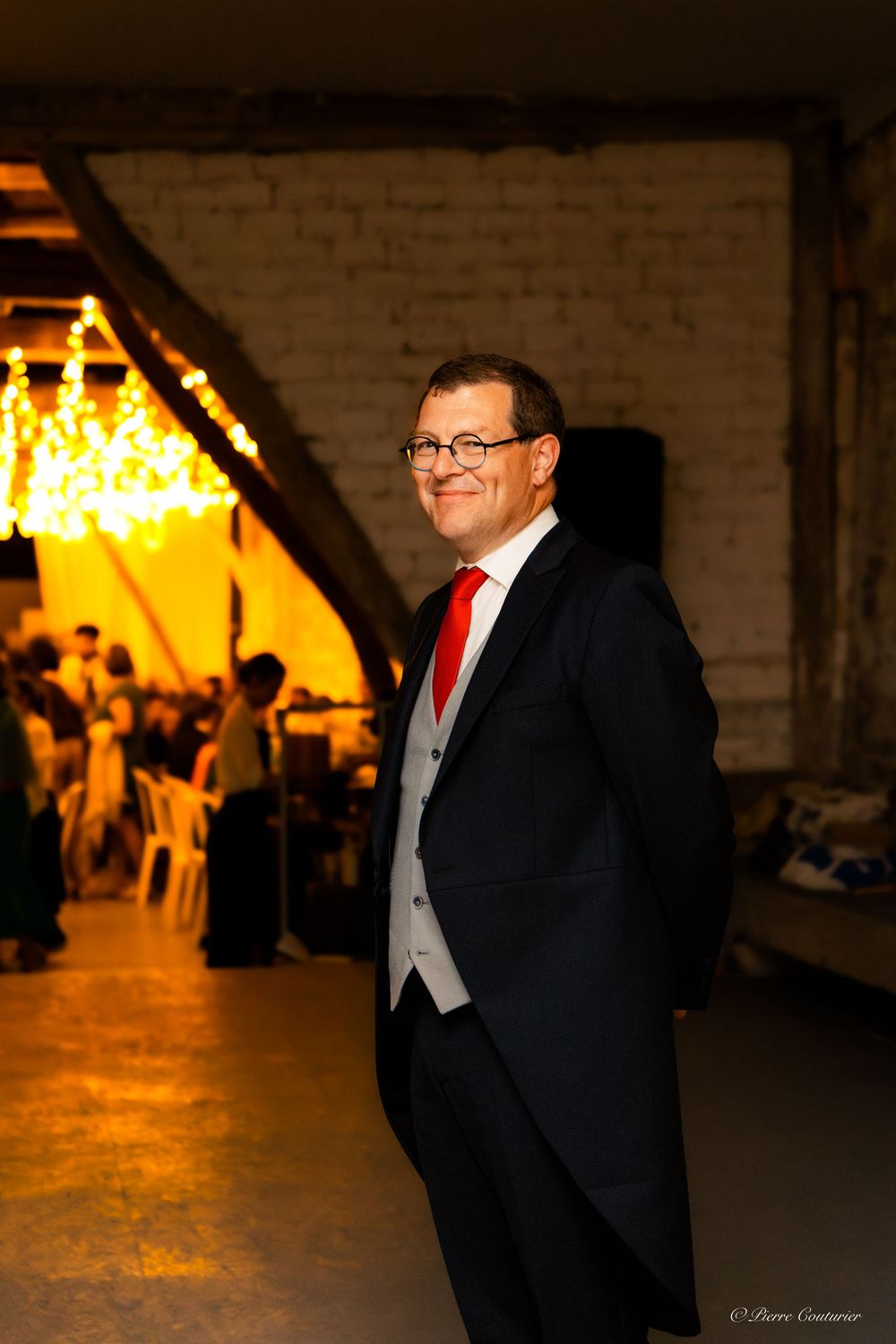 A smiling man in a black suit with red tie poses confidently at an elegant wedding reception venue with warm lighting.