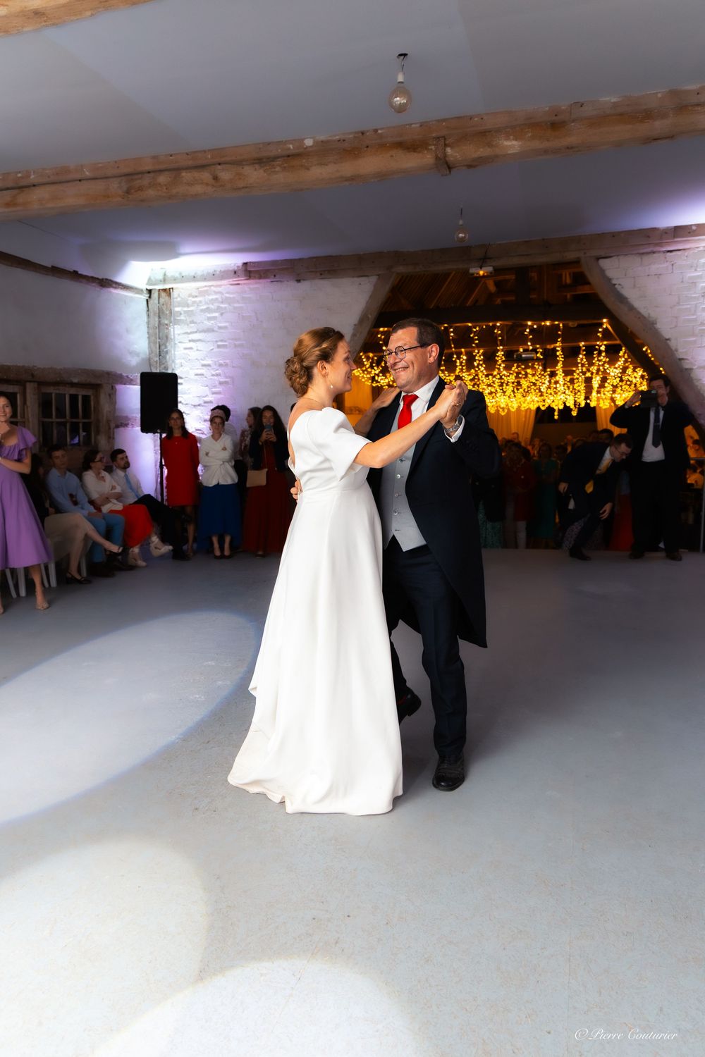 Bride and groom share their first dance in a rustic barn venue with fairy lights and guests watching.