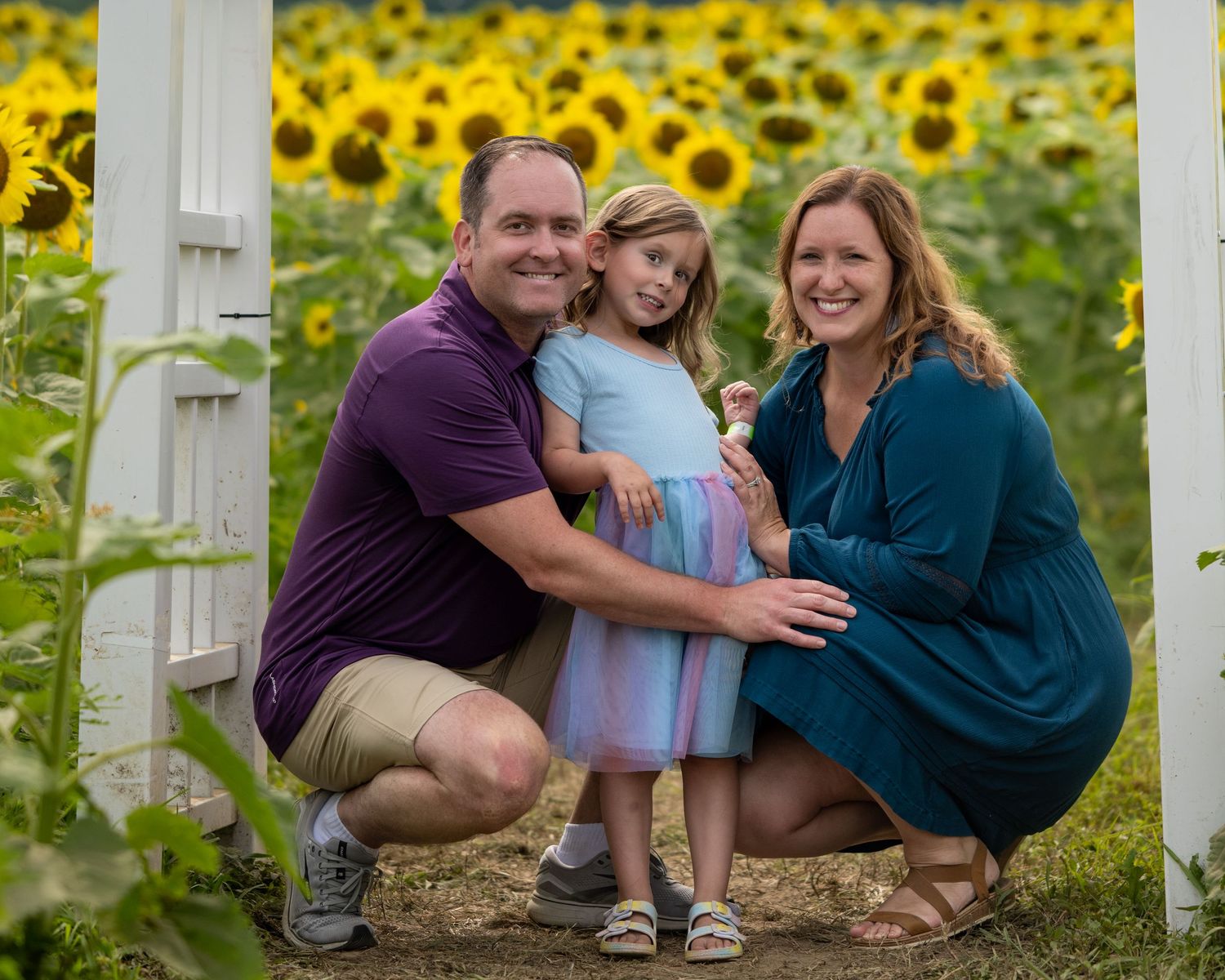 Family of three smiling in a sunflower field, parents crouching beside young girl in blue dress.