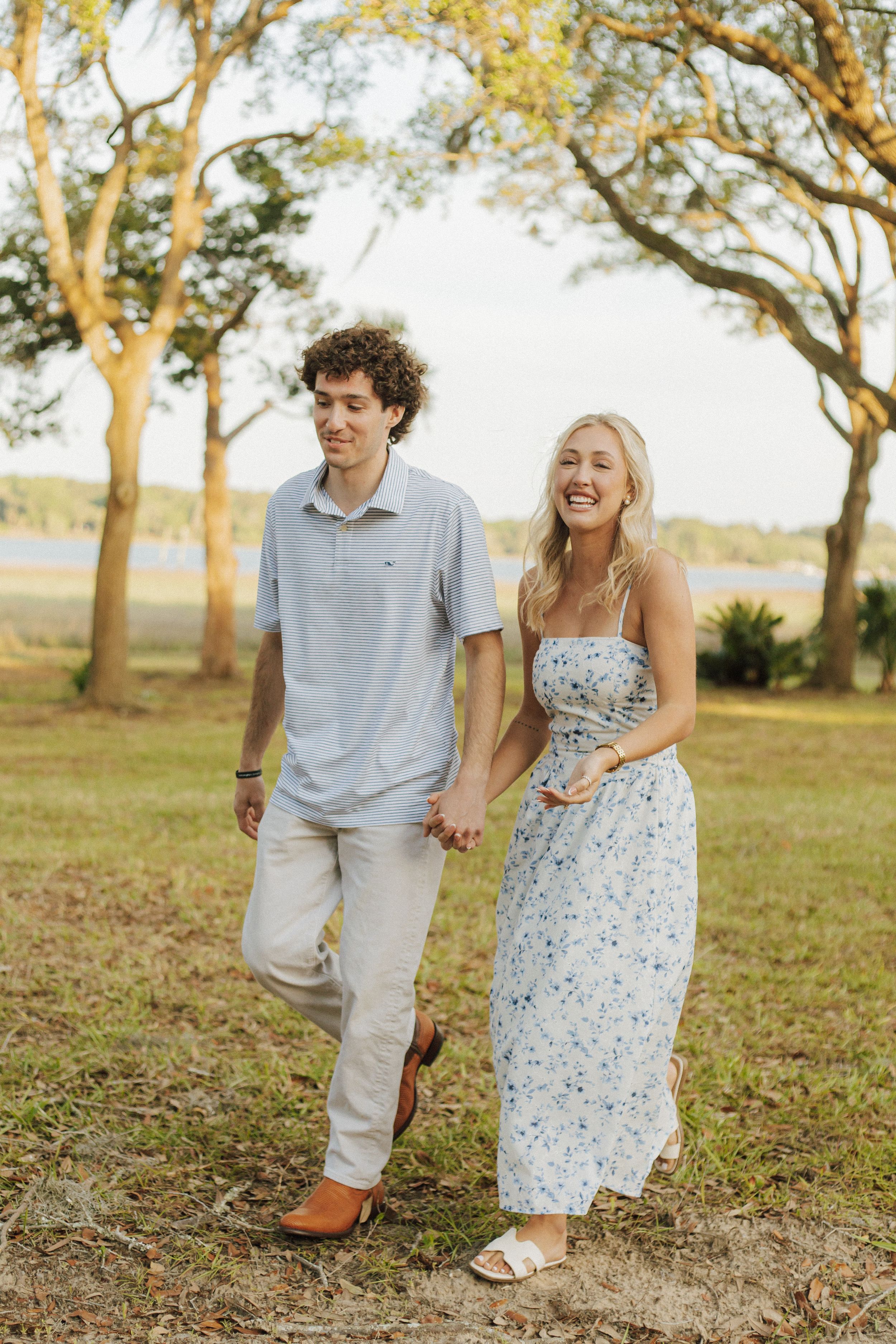 A smiling couple holds hands while walking outdoors near trees and a lake, dressed in casual summer attire.