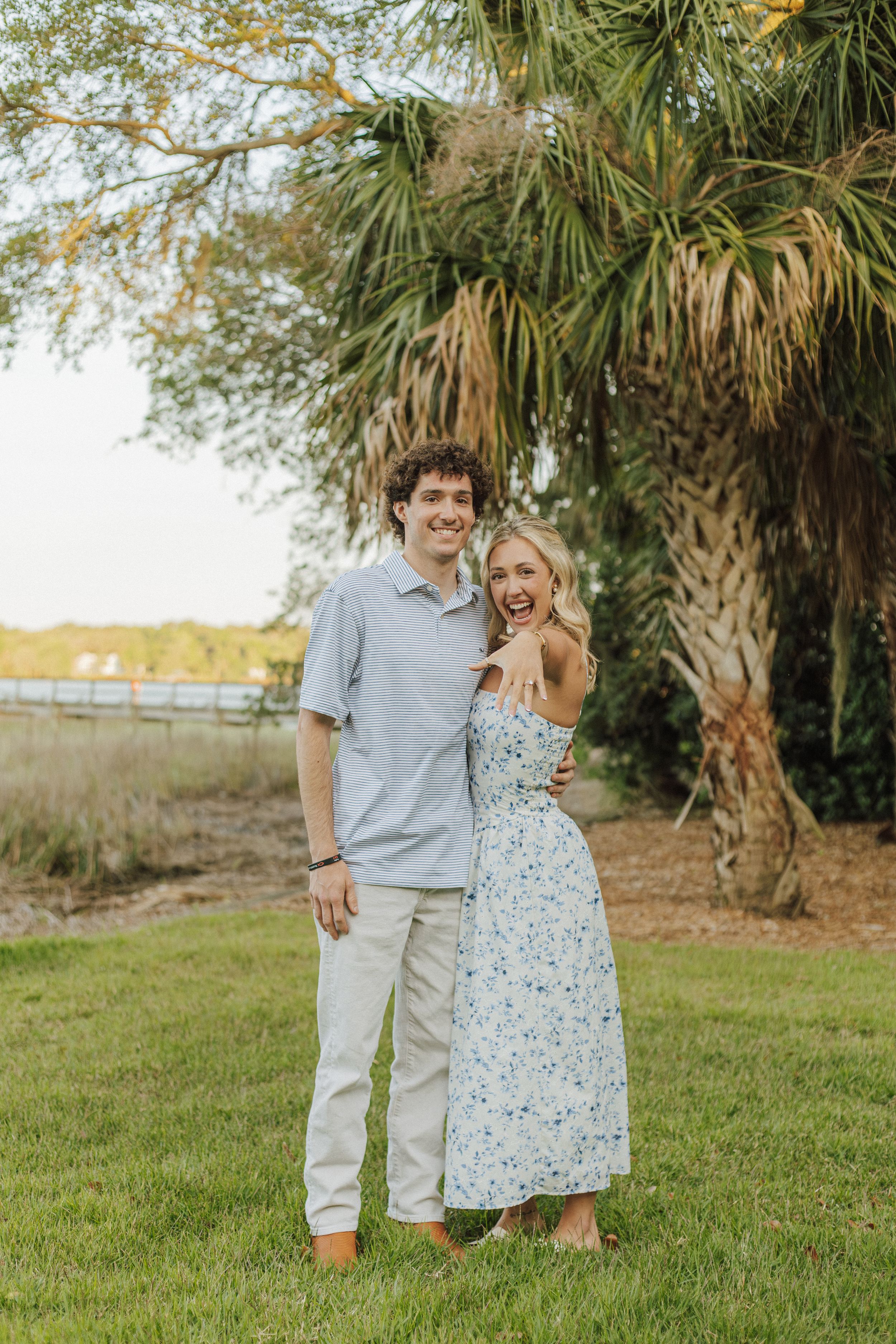 A smiling couple poses together on green grass beneath a palm tree in a sunny outdoor setting.