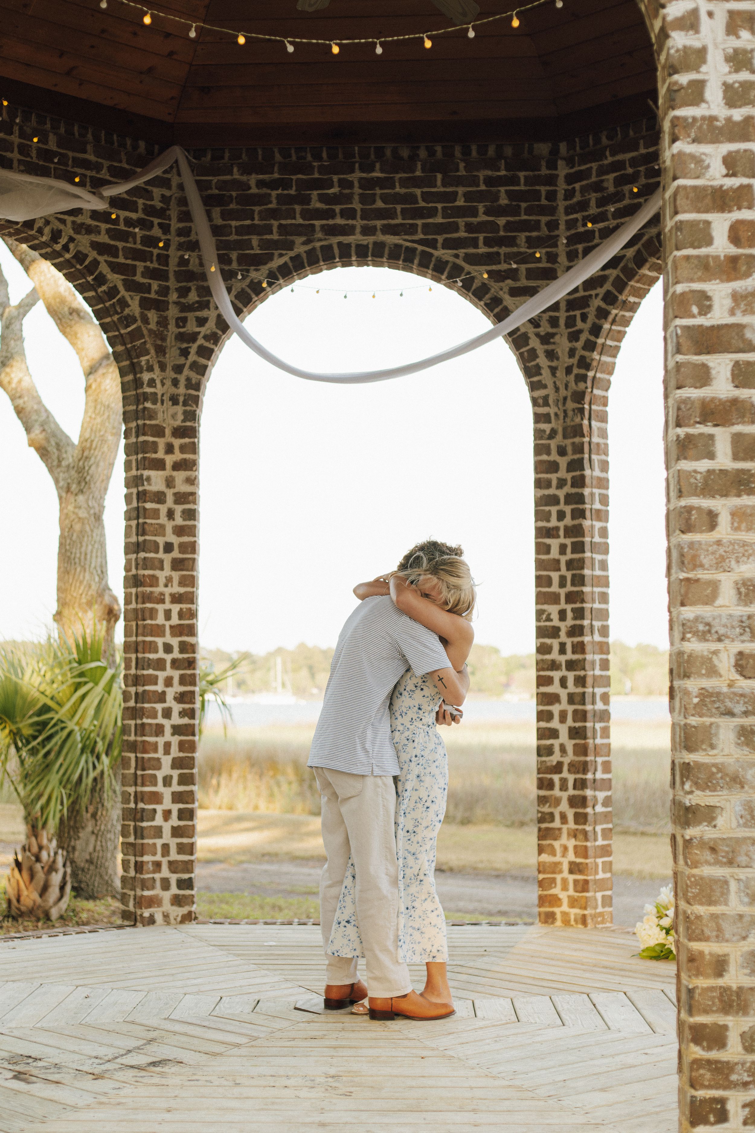 Couple embracing under brick arched pavilion with string lights, overlooking golden marsh landscape at sunset.