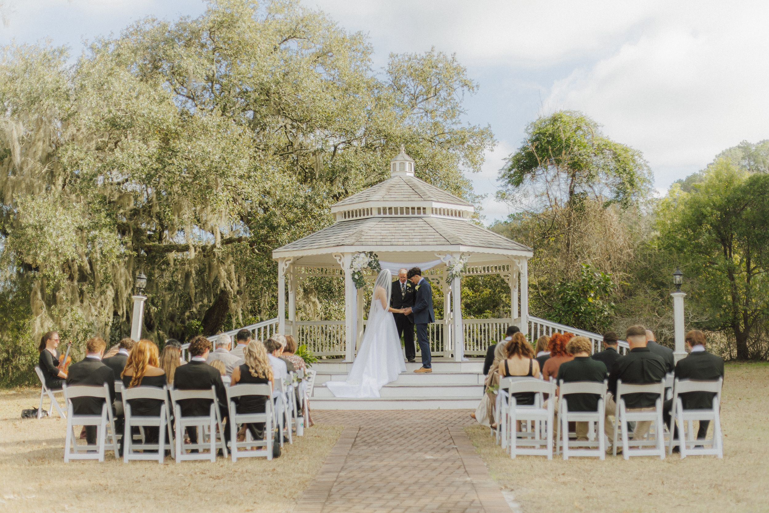 Outdoor wedding ceremony under a white gazebo with guests seated in white chairs surrounded by lush green trees.