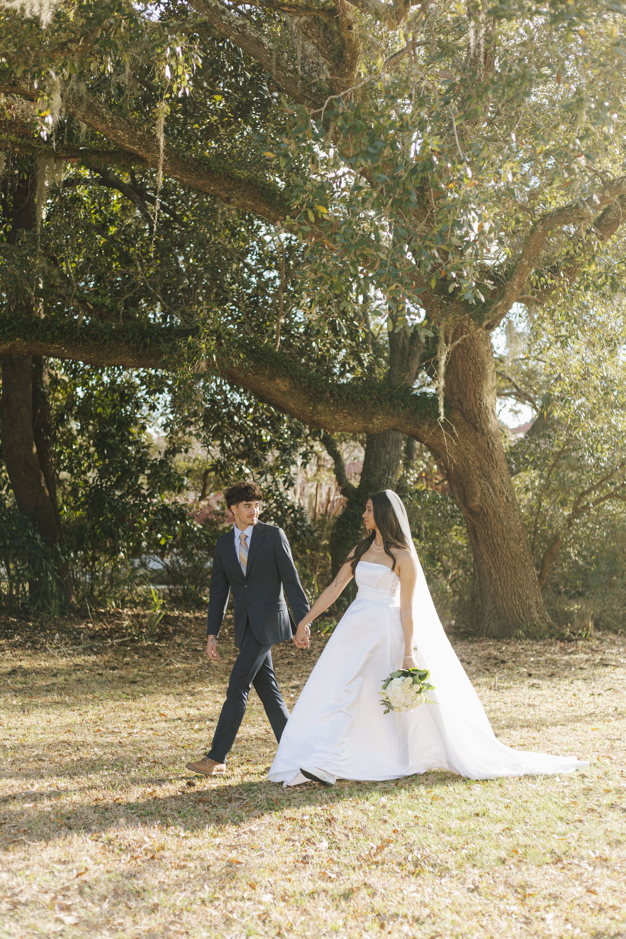 A bride and groom walk hand-in-hand beneath sprawling oak trees on their wedding day outdoors.