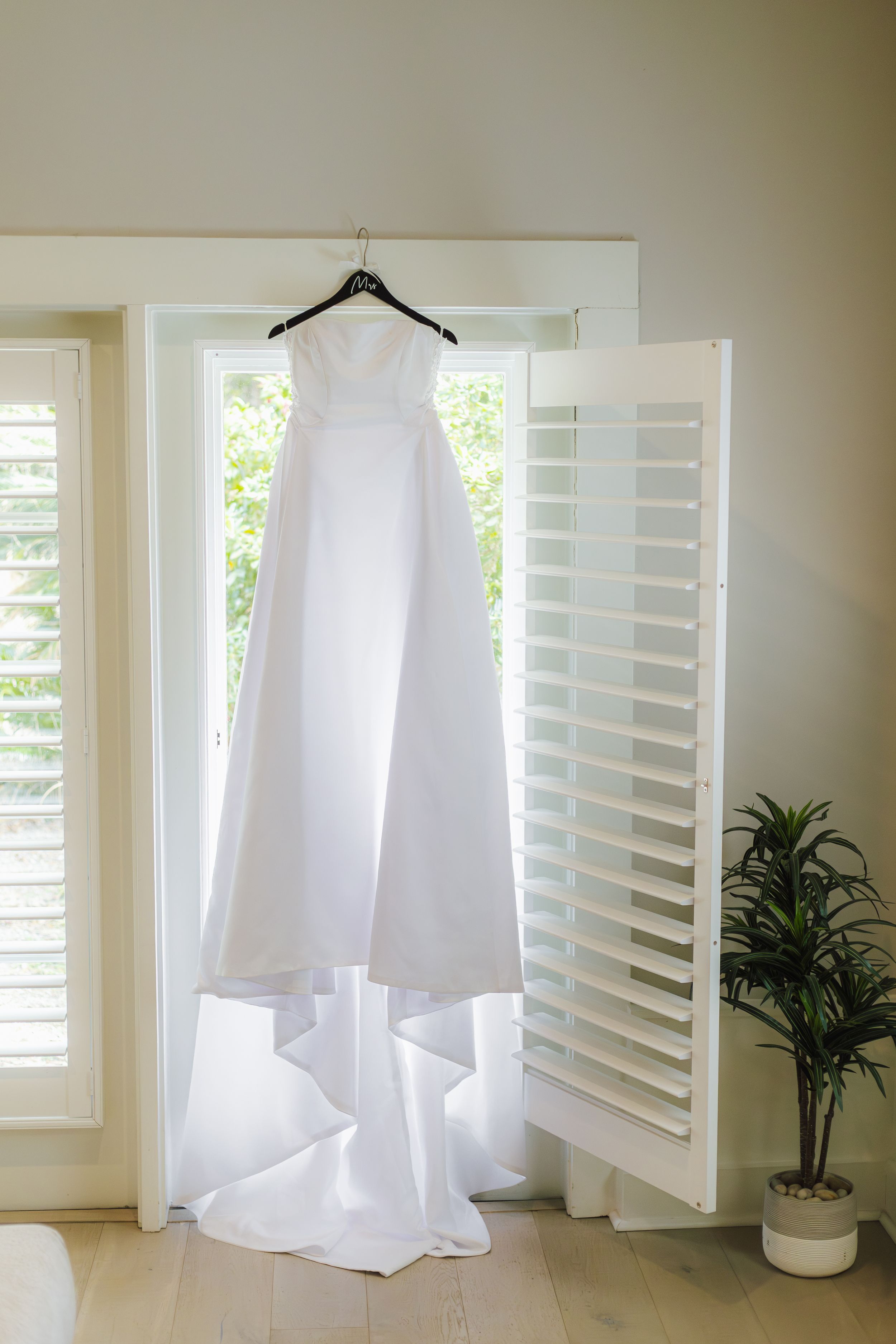 Elegant white wedding dress hanging on a black hanger in a bright room with plantation shutters and a potted plant.