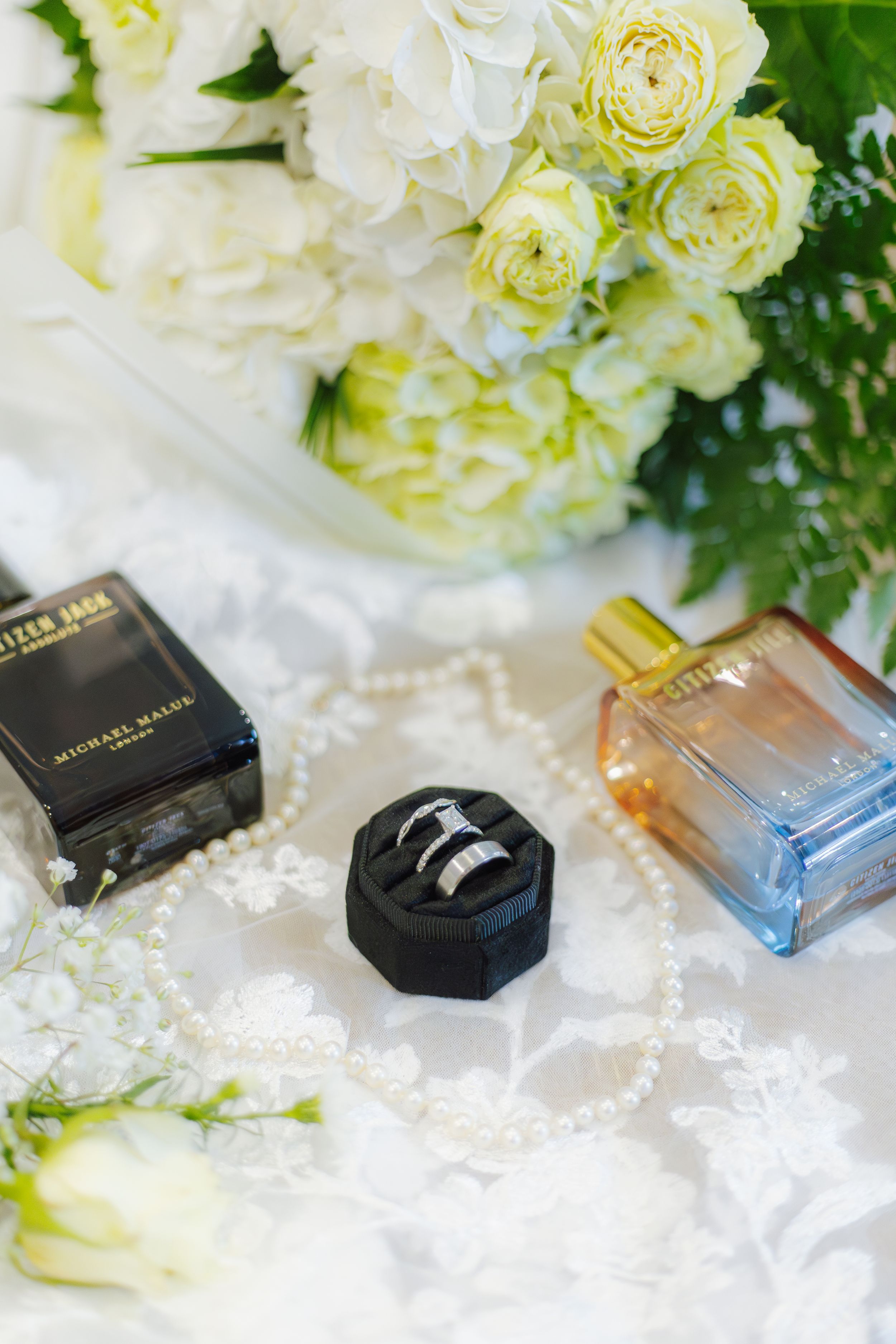 Wedding rings in black velvet box surrounded by pearl necklace, perfume bottles, and white floral bouquet on lace.