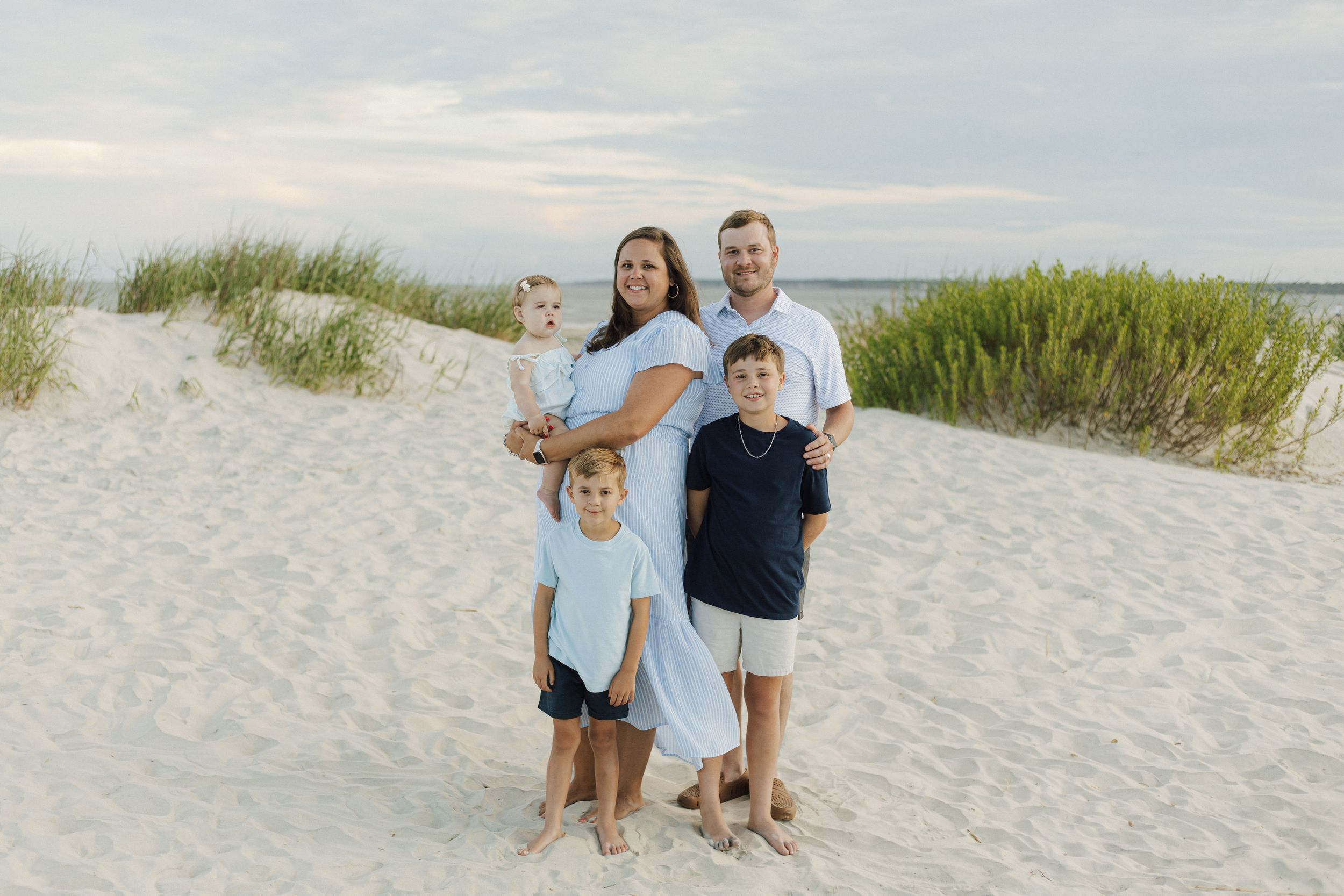Family of five posing together on a sandy beach with sea grass and calm water in the background.