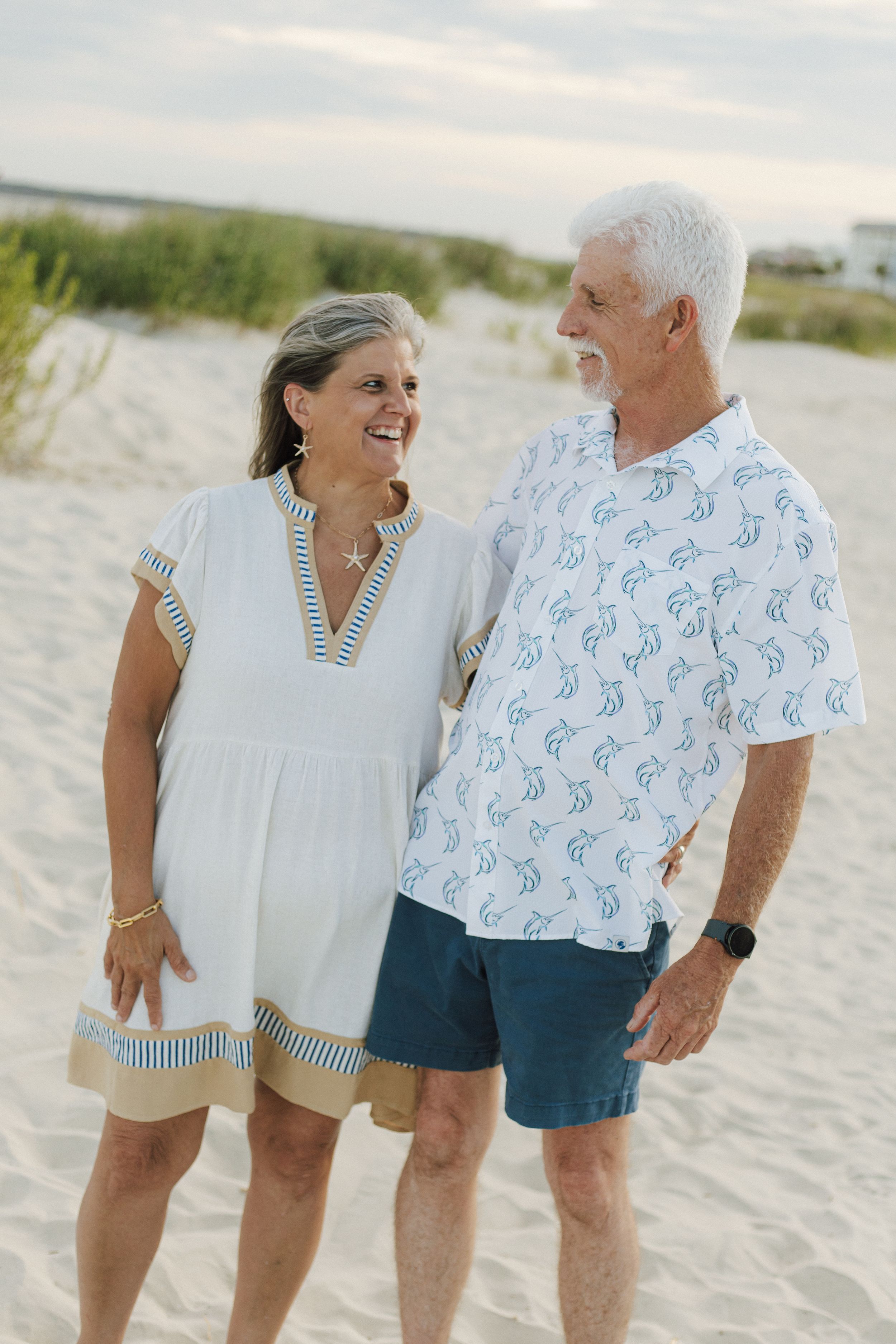 Happy senior couple smiling at each other on sandy beach, wearing casual summer outfits in white and blue.