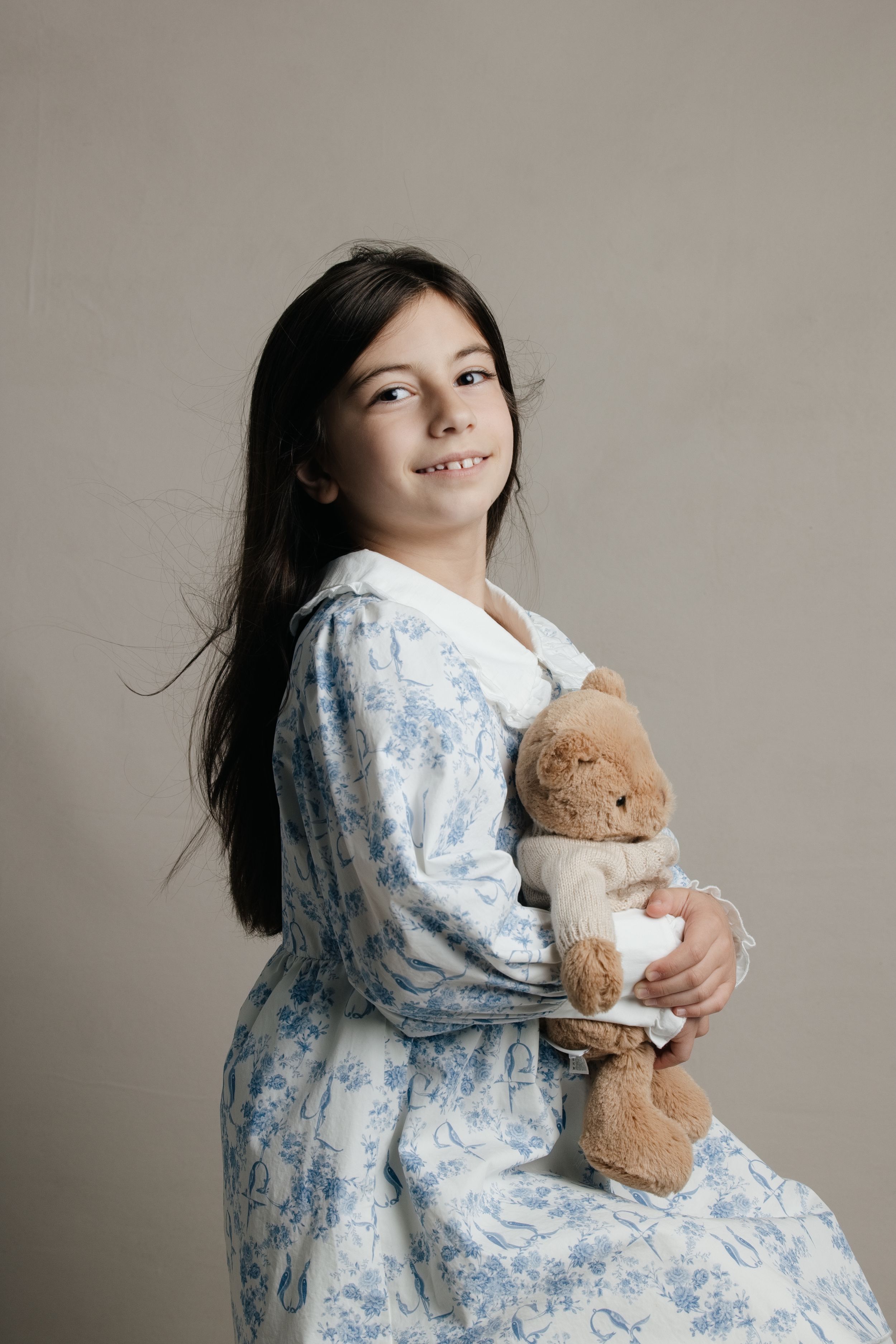 Young girl in blue floral dress hugging a teddy bear, smiling over her shoulder against a neutral background.