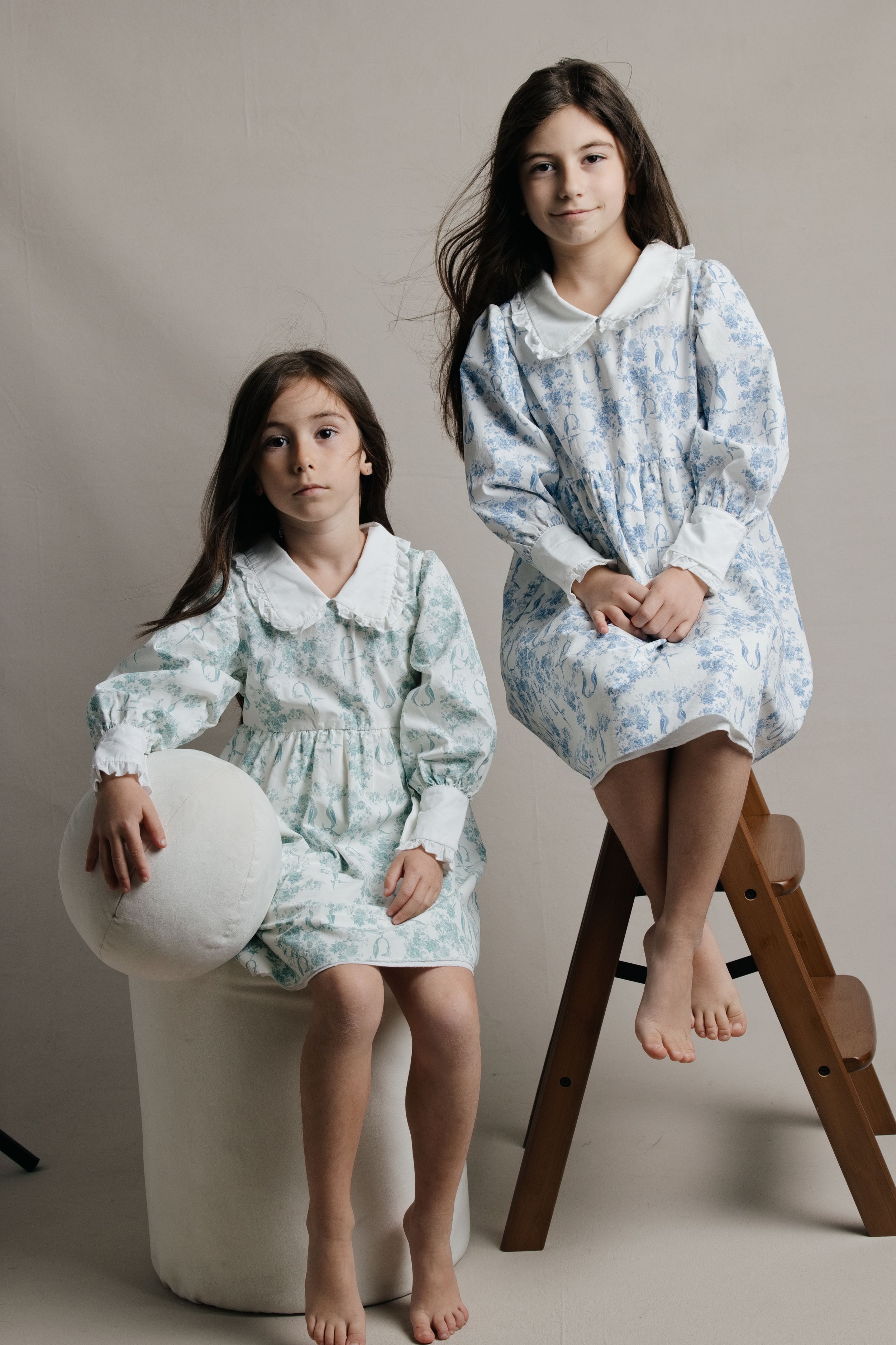 Two girls in matching floral dresses with white collars pose in a studio, one seated on a stool, one on a wooden ladder.